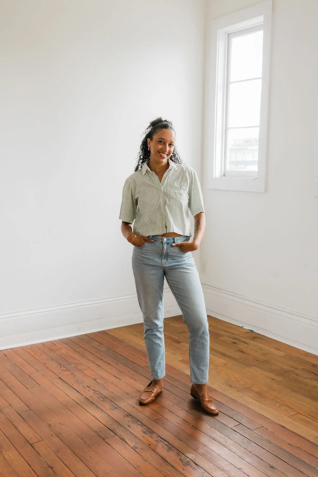 A young woman smiling in a bright, empty room with hardwood floors and a large window with natural light.