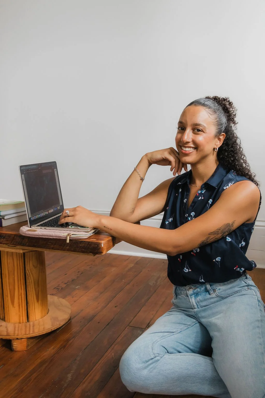 A woman with curly hair tied up in a ponytail, wearing a sleeveless floral blouse and light jeans, smiling while sitting on her knees at a wooden table with a laptop and some books in a room with wooden floor and plain white wall.
