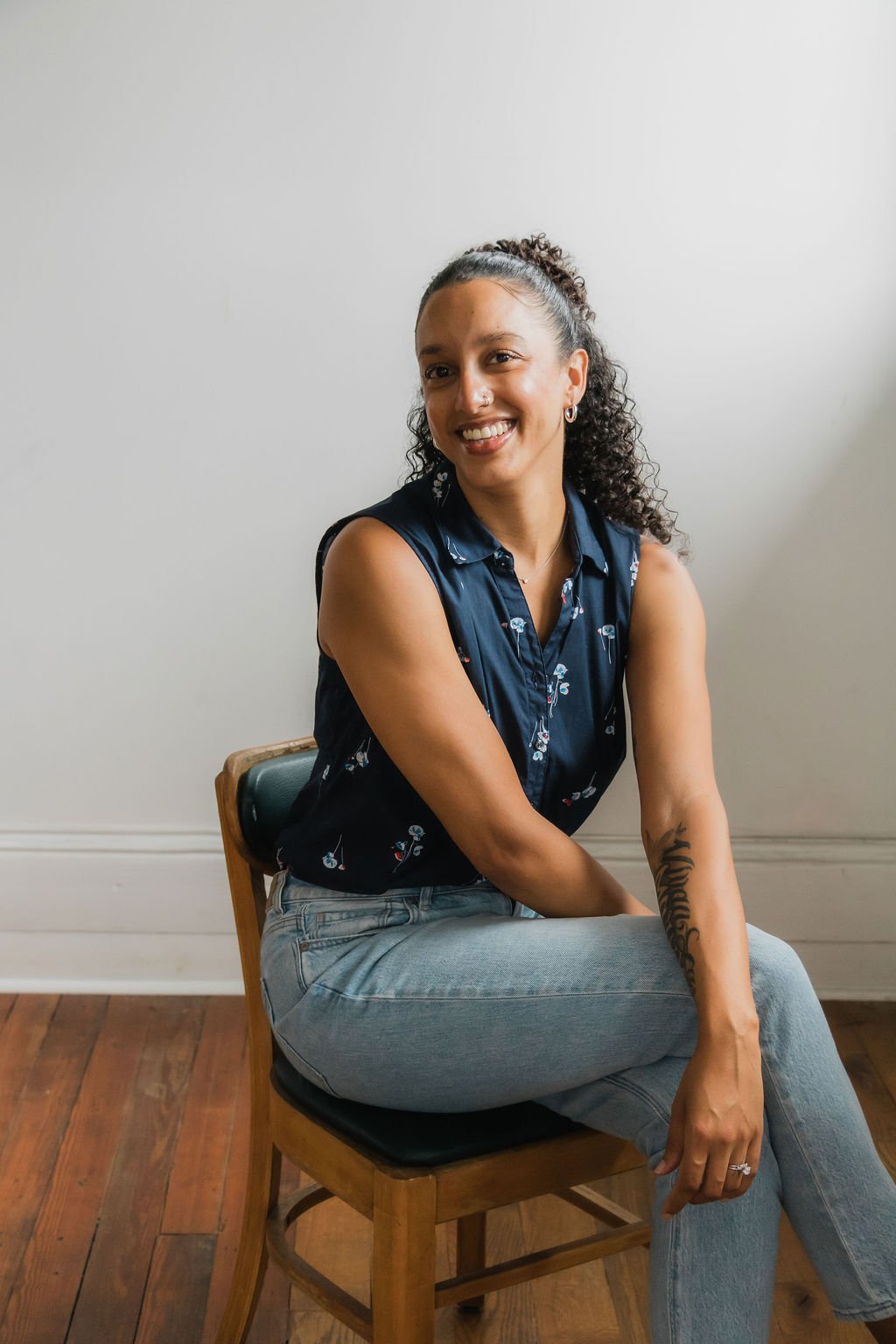 A woman with curly hair tied back, wearing a sleeveless dark blue floral top and light blue jeans, sitting on a wooden chair with a black cushion, smiling at the camera against a plain white wall background.