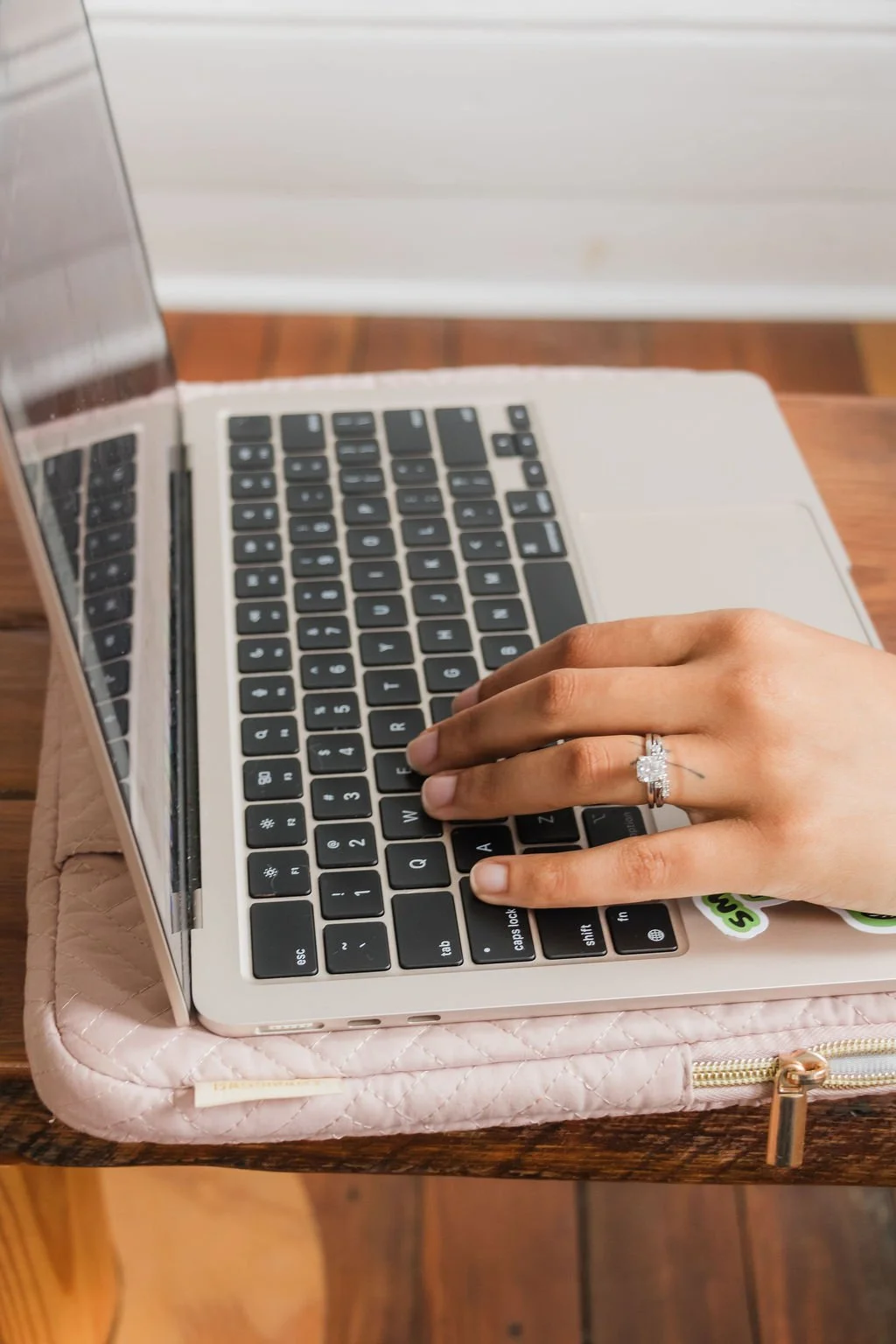 A person's hand with a diamond ring on the ring finger typing on a laptop keyboard. The laptop is placed on a Pink quilted laptop sleeve, on a wooden table.