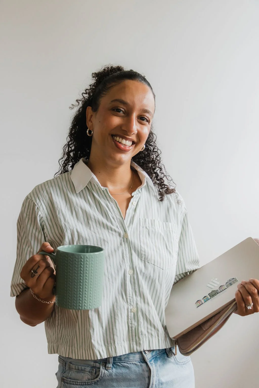 A woman with curly hair smiling, wearing a striped button-up shirt, holding a blue mug in one hand and a notebook in the other, standing against a plain white wall.