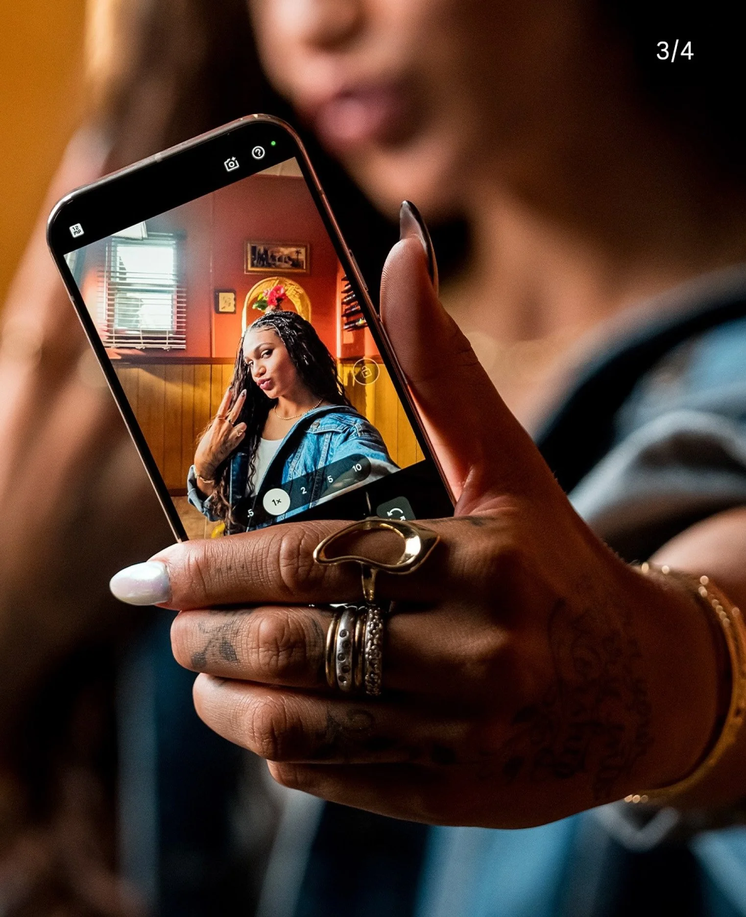 Still from commercial set produced by Daedalus LLC. Close-up of a person's hand holding a smartphone, with a photo of a woman making a peace sign on the screen, in a room with wooden and red walls, window, and framed pictures.