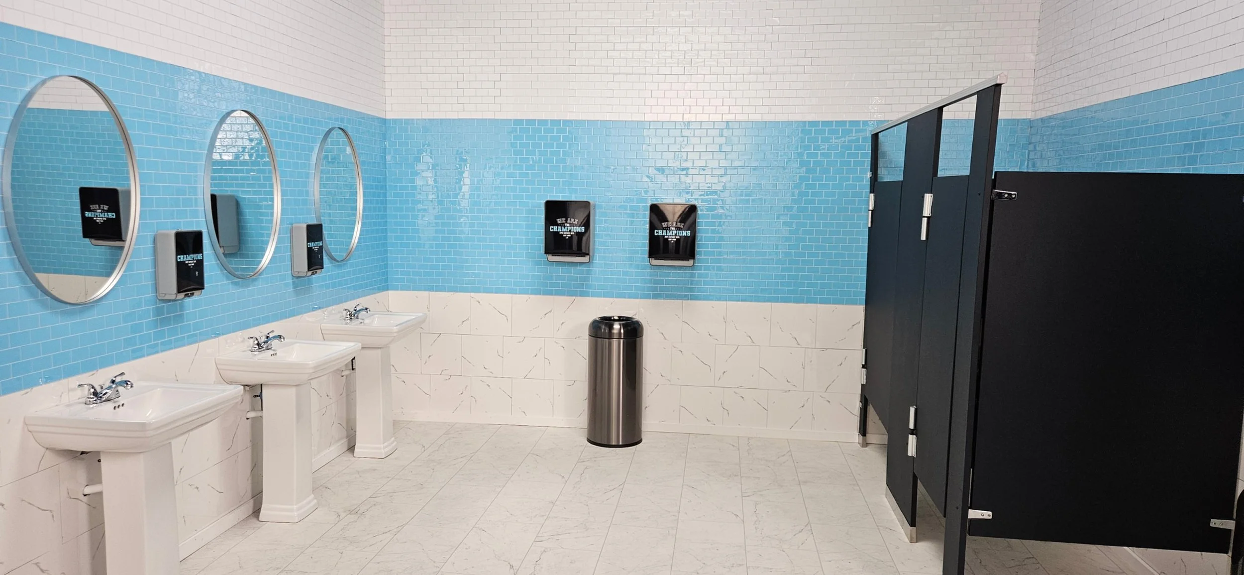 Custom built set on soundstage made by Daedallus LLC as part of the Art Department. A clean men's restroom with three white sinks, three mirrors, and two paper towel dispensers on a blue tiled wall. 