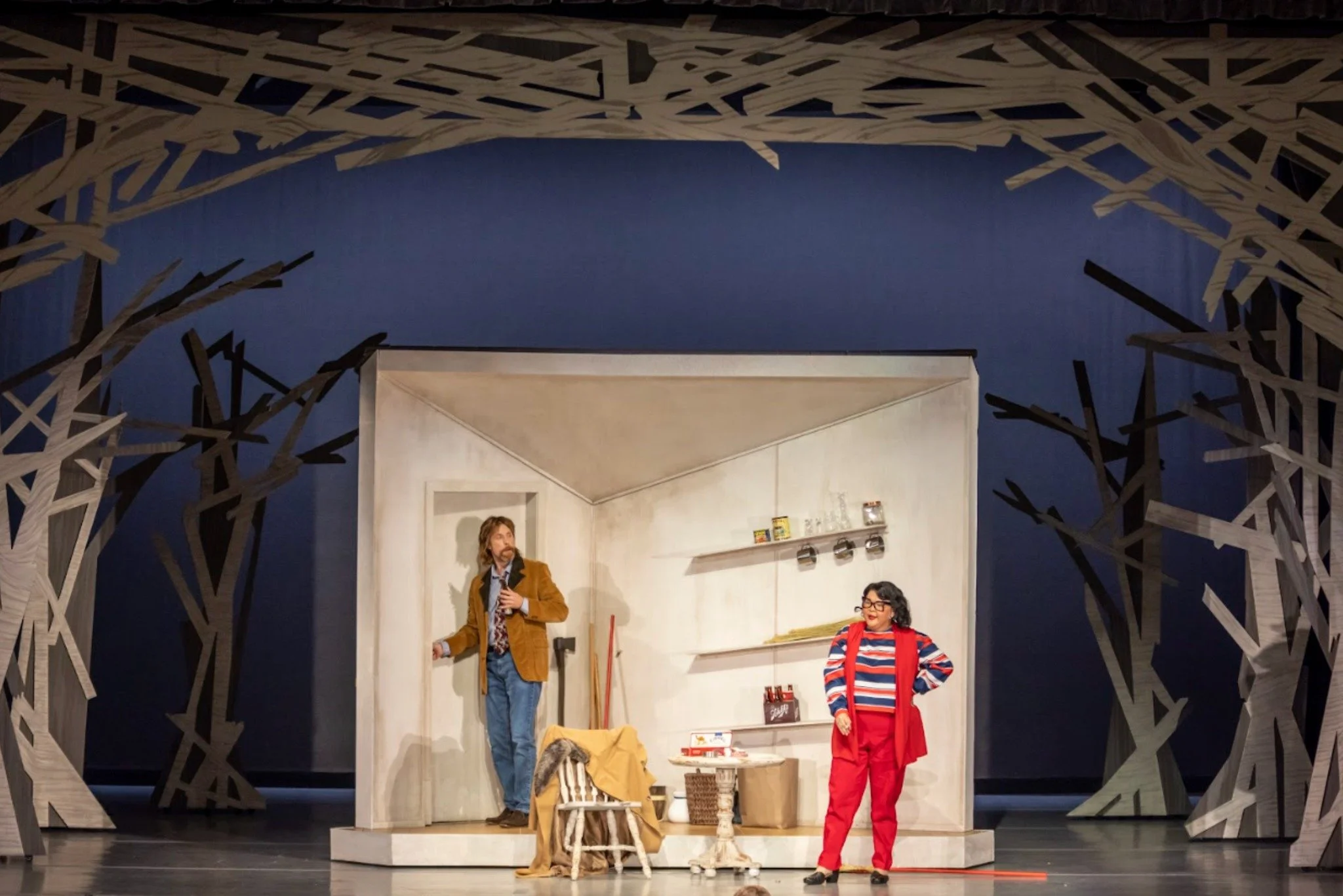 The image shows a theater stage set with a rustic, white wooden house and an arched, wooden lattice frame resembling tree branches framing the scene. There are two actors, a man and a woman, engaged in a conversation. The man has long hair and a bear