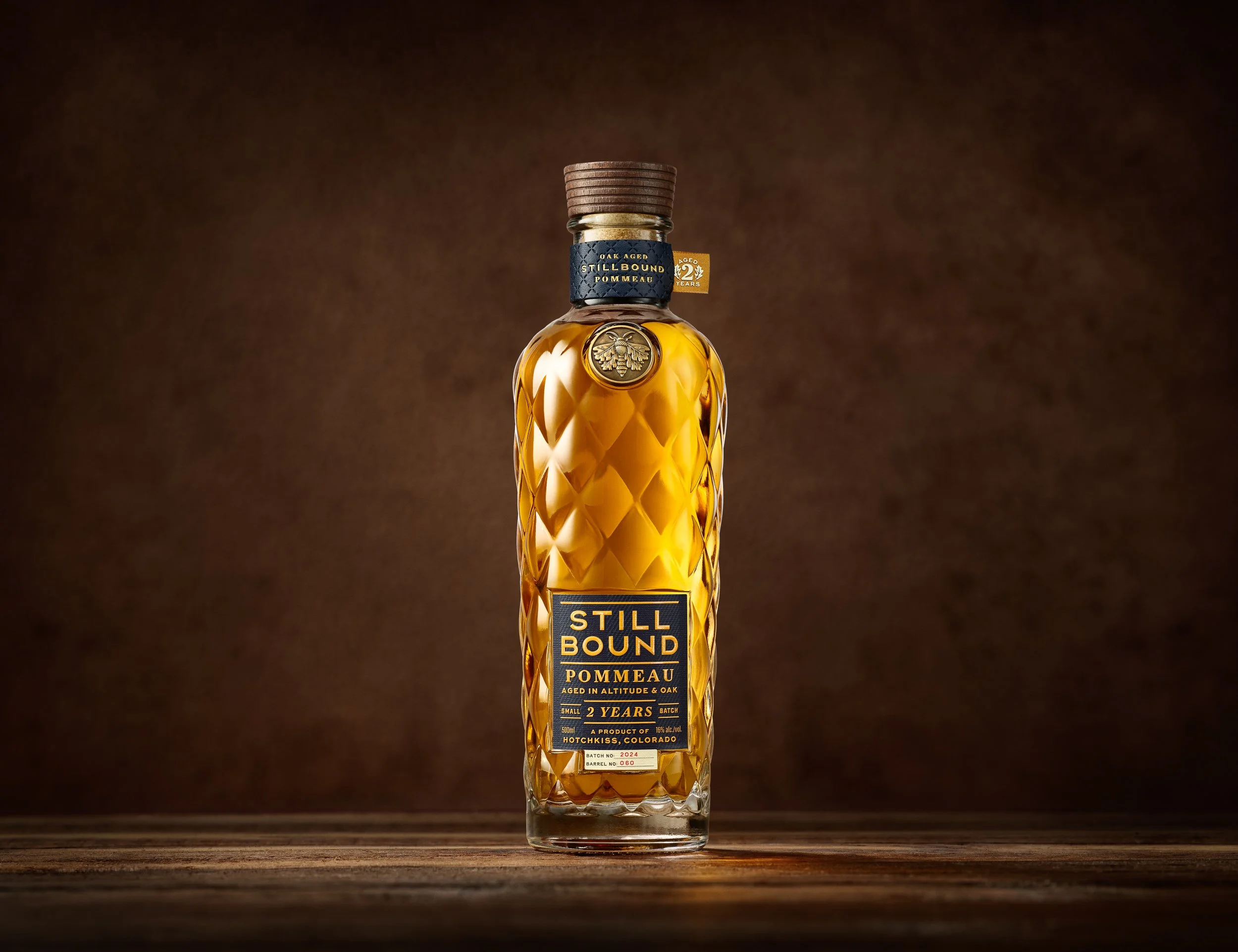 A glass bottle of Still Bound Pommeau, a type of alcoholic beverage, with a wooden cap, gold medallion, and detailed textured glass design, set against a dark brown background.