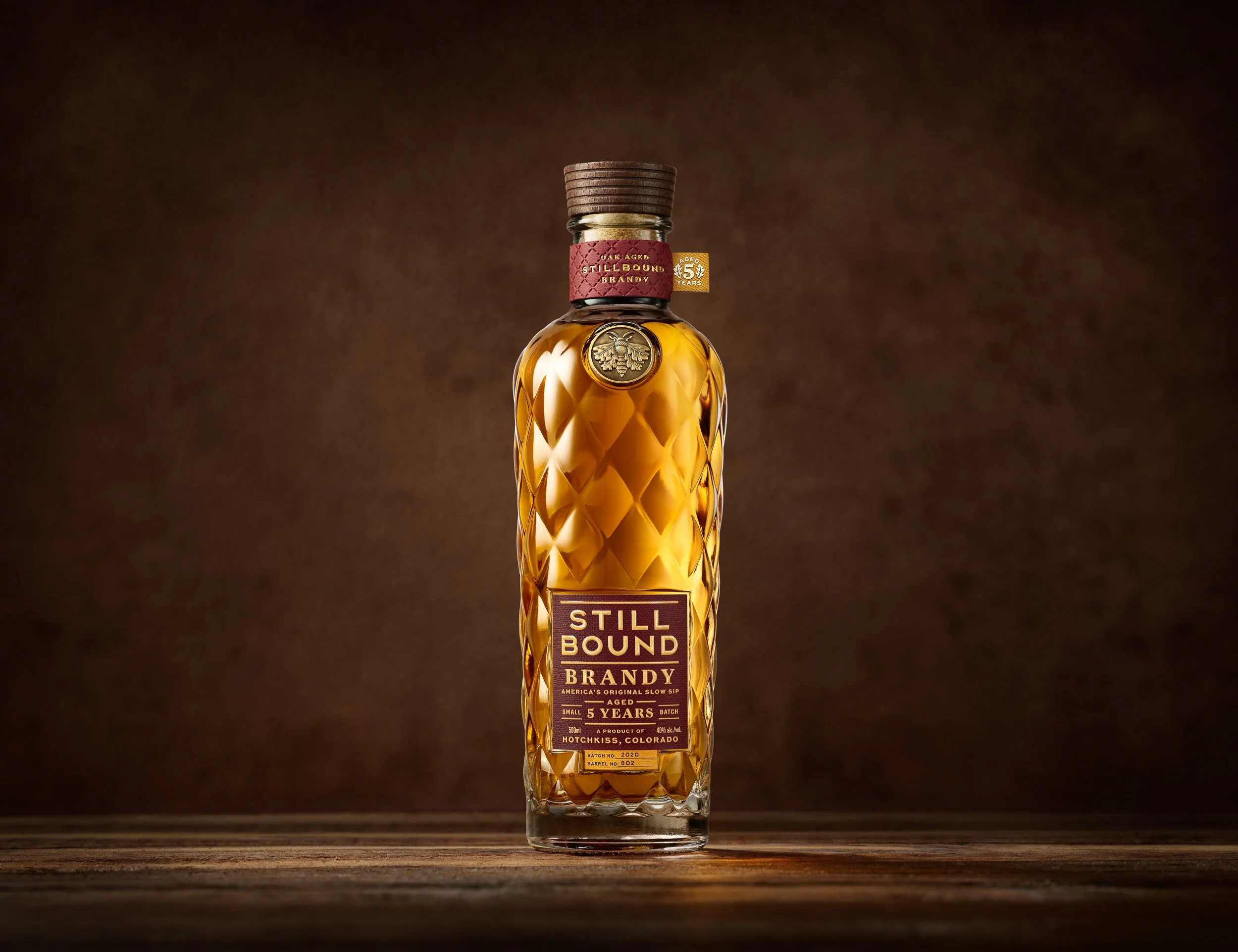 A tall, clear glass bottle of Still Bound Brandy with a textured diamond pattern on the glass, a gold medallion emblem on the front, a red label wrapping around the neck with gold text, and a wooden cap, placed on a wooden surface against a dark brown background.