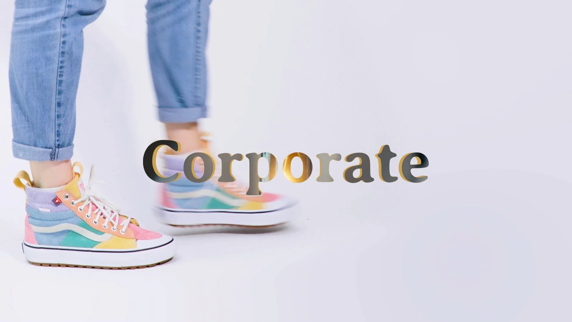 Person wearing colorful rainbow sneakers and rolled-up jeans, standing on a skateboard with the word "Corporate" overlapping the image on a white background.