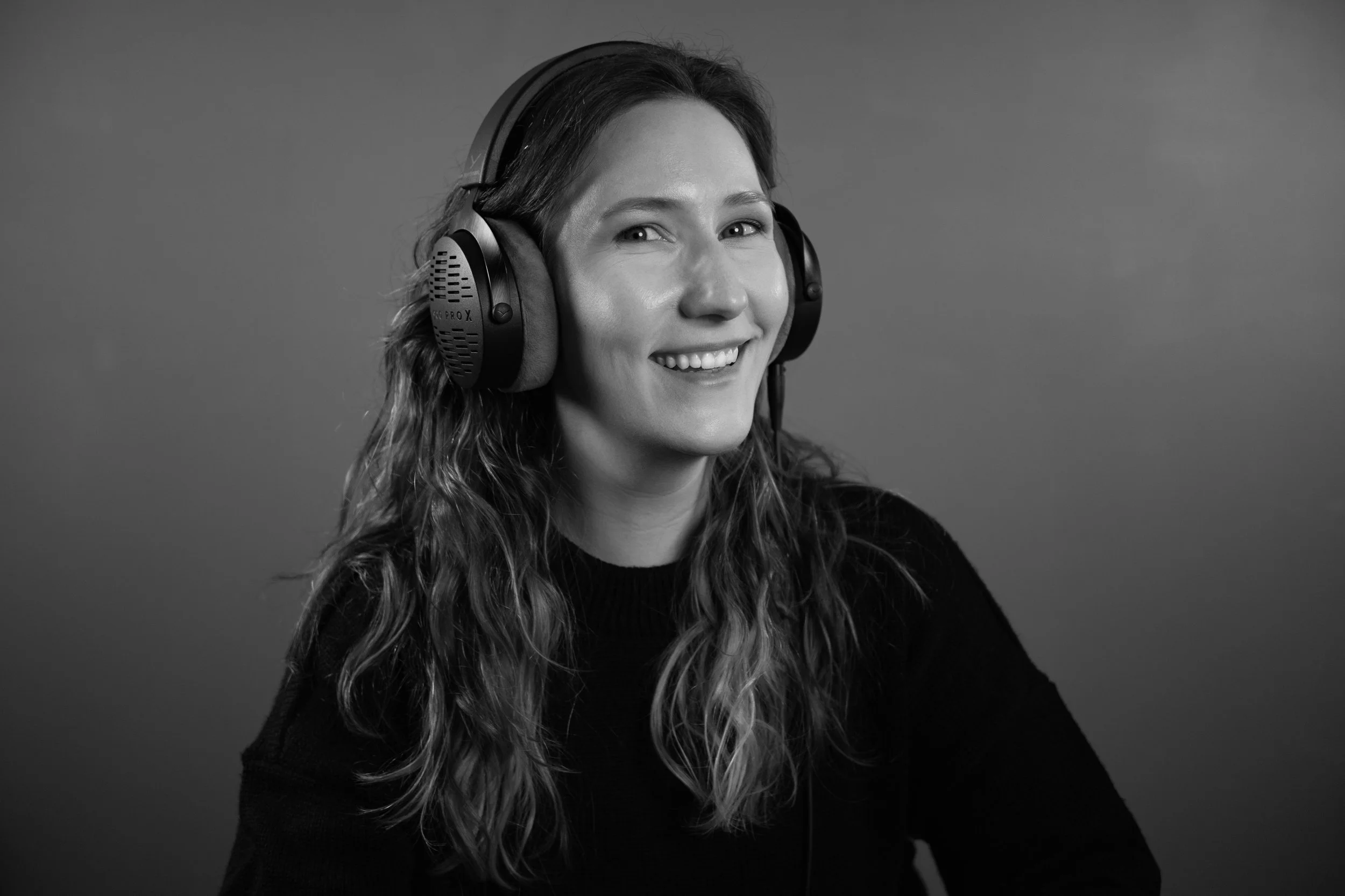 Black and white photo of a smiling woman with long, wavy hair wearing large headphones, against a plain background.