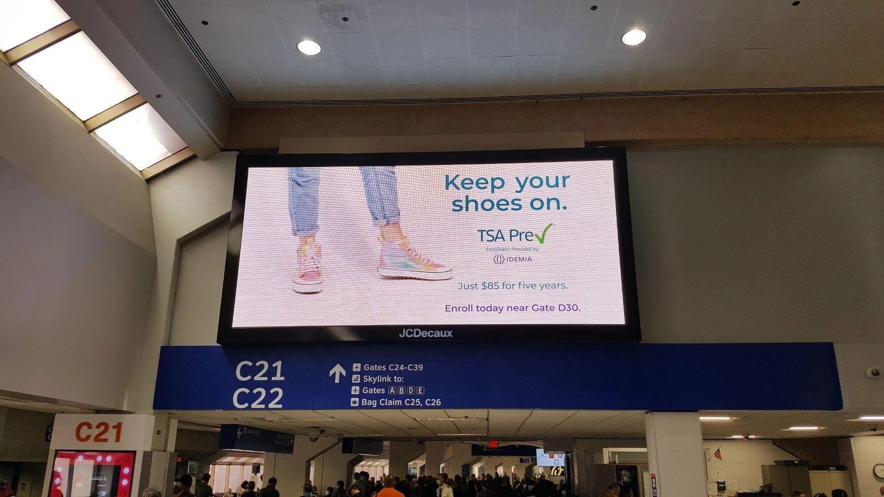 An airport information screen with an advertisement that says "Keep your shoes on" and promotes TSA Pre✓ enrollment. The ad features an illustration of a person wearing sneakers and jeans. Below, there are gate signs and directions to gates, skylink, and baggage claim.