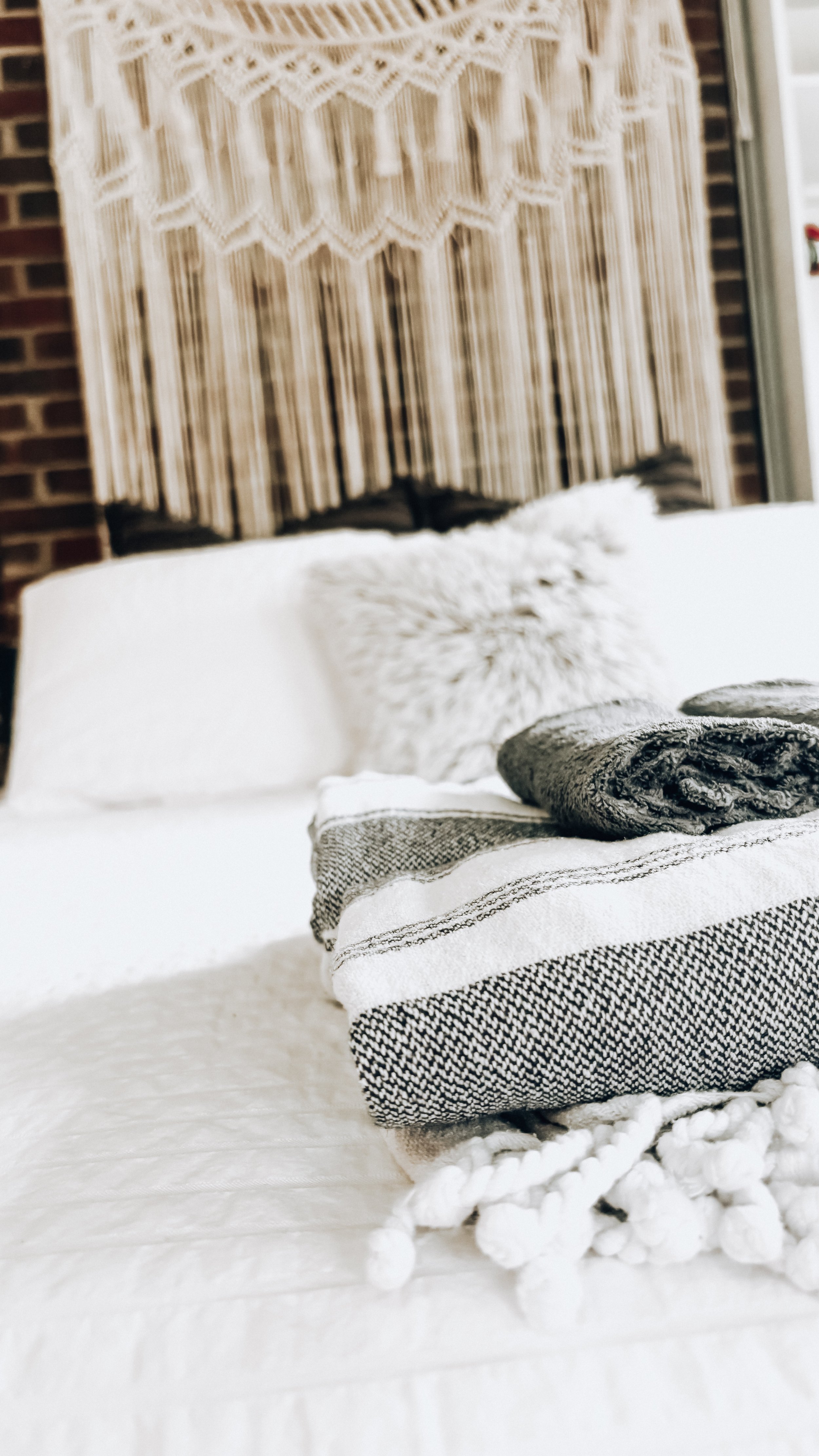 Bedroom with a bed, pillows, and blankets in neutral tones; textile wall hanging above the bed; exposed brick wall on the side; window on the right.