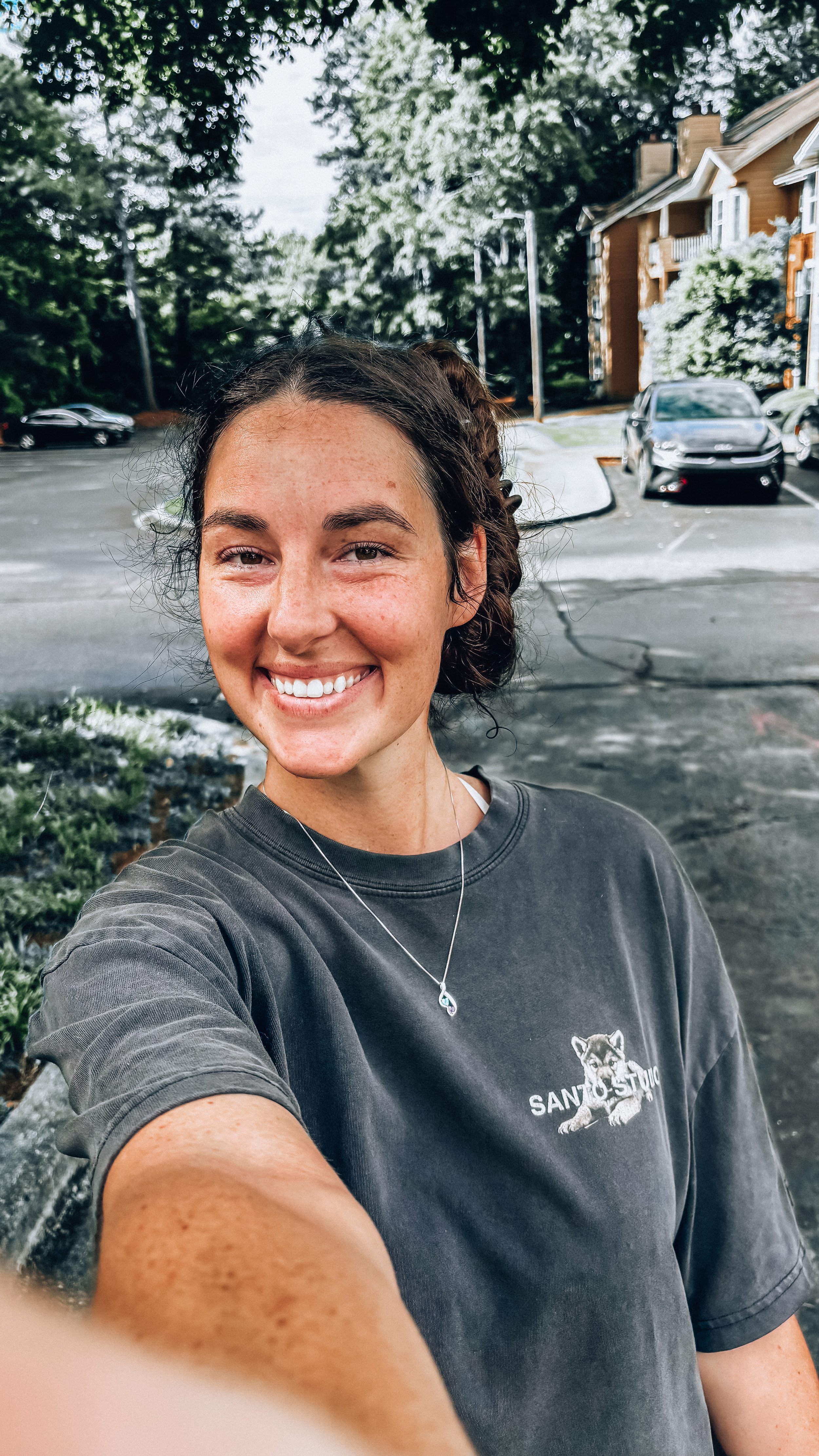 A woman taking a selfie outdoors with trees, parked cars, and residential buildings in the background. She is smiling, wearing a dark T-shirt with a wolf graphic and a necklace.