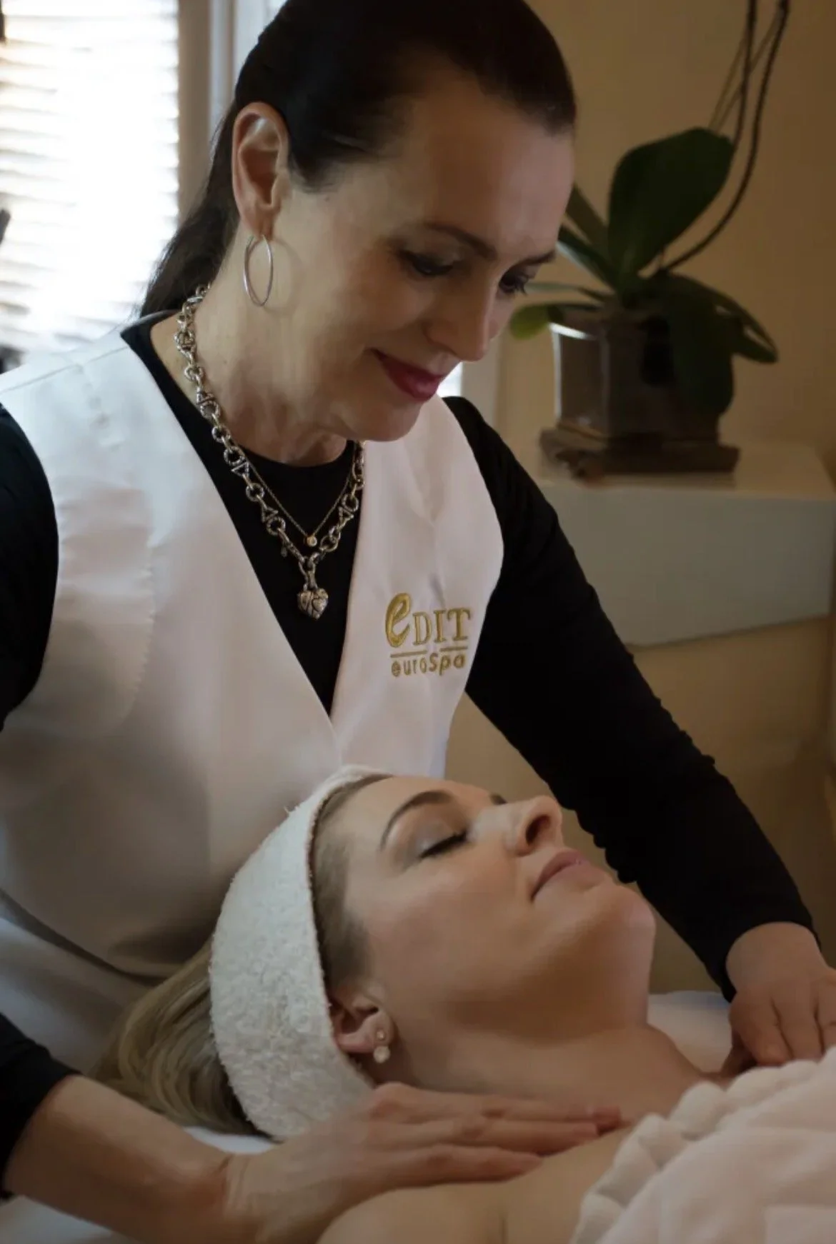 A woman with dark hair doing a facial treatment on a woman lying down with her eyes closed, in a spa setting with a potted plant in the background.