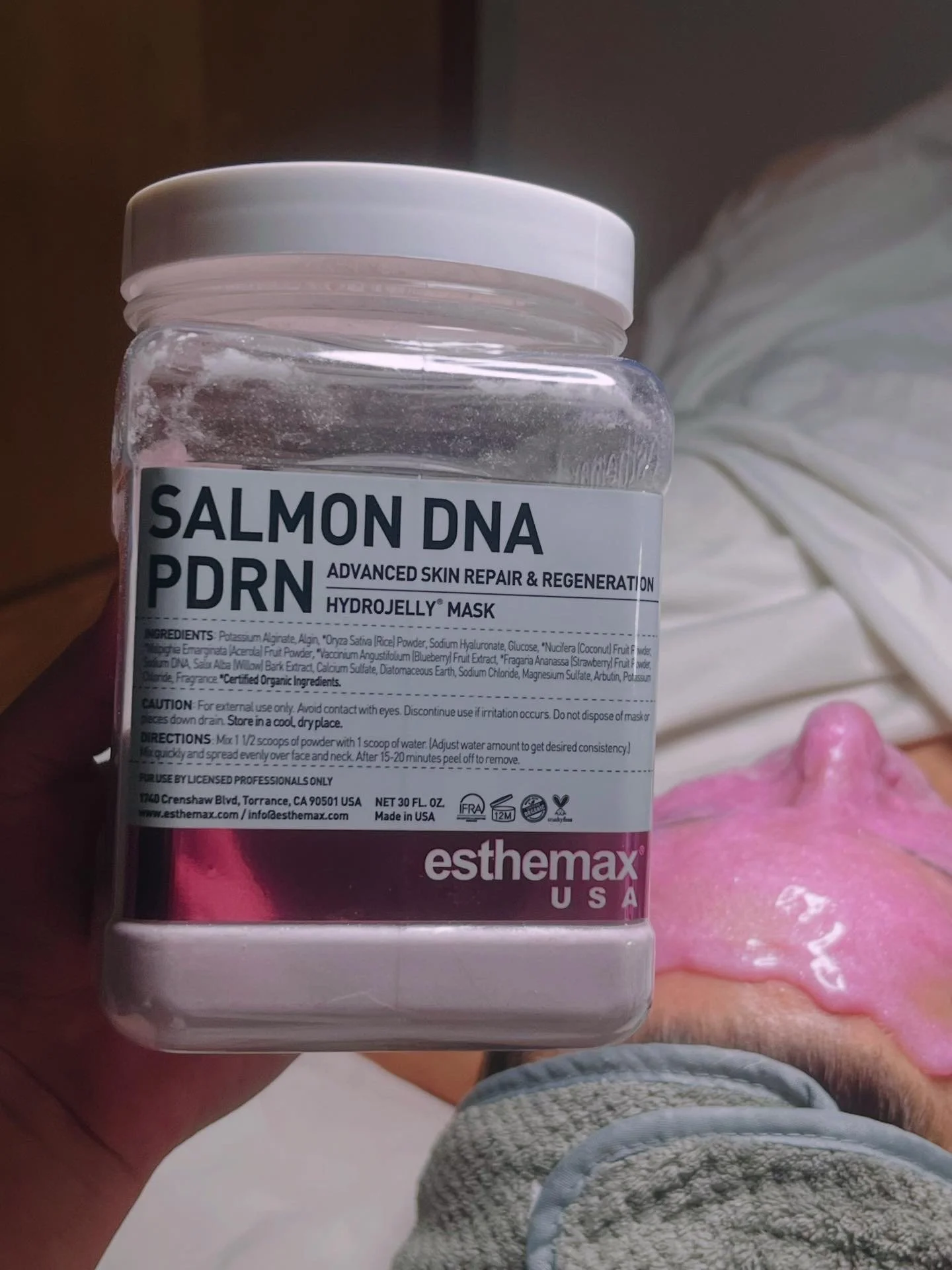 A clear plastic jar of Esthemax Salmon DNA PDRN Hydrojelly Mask held in a person's hand, with a blurred background and part of a person's leg and a pink object visible.