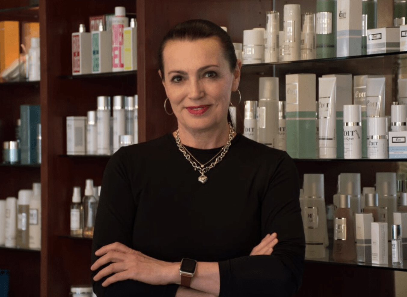 A woman with dark hair, wearing a black top, silver jewelry, and a smartwatch, standing in front of shelves filled with skincare products.