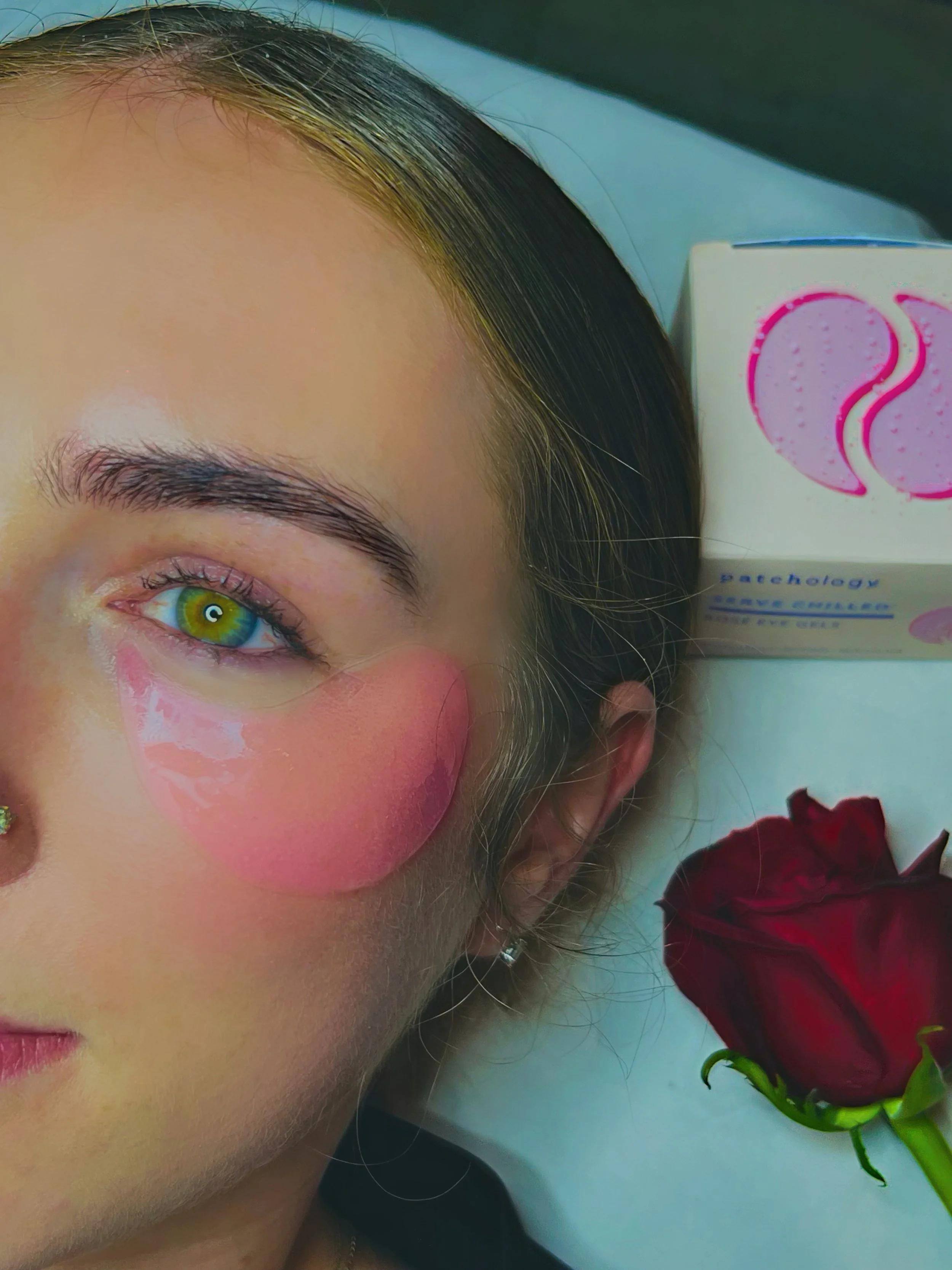 Close-up of a woman with green eyes, wearing pink blush and eye makeup, and a red rose on the right side of the image.