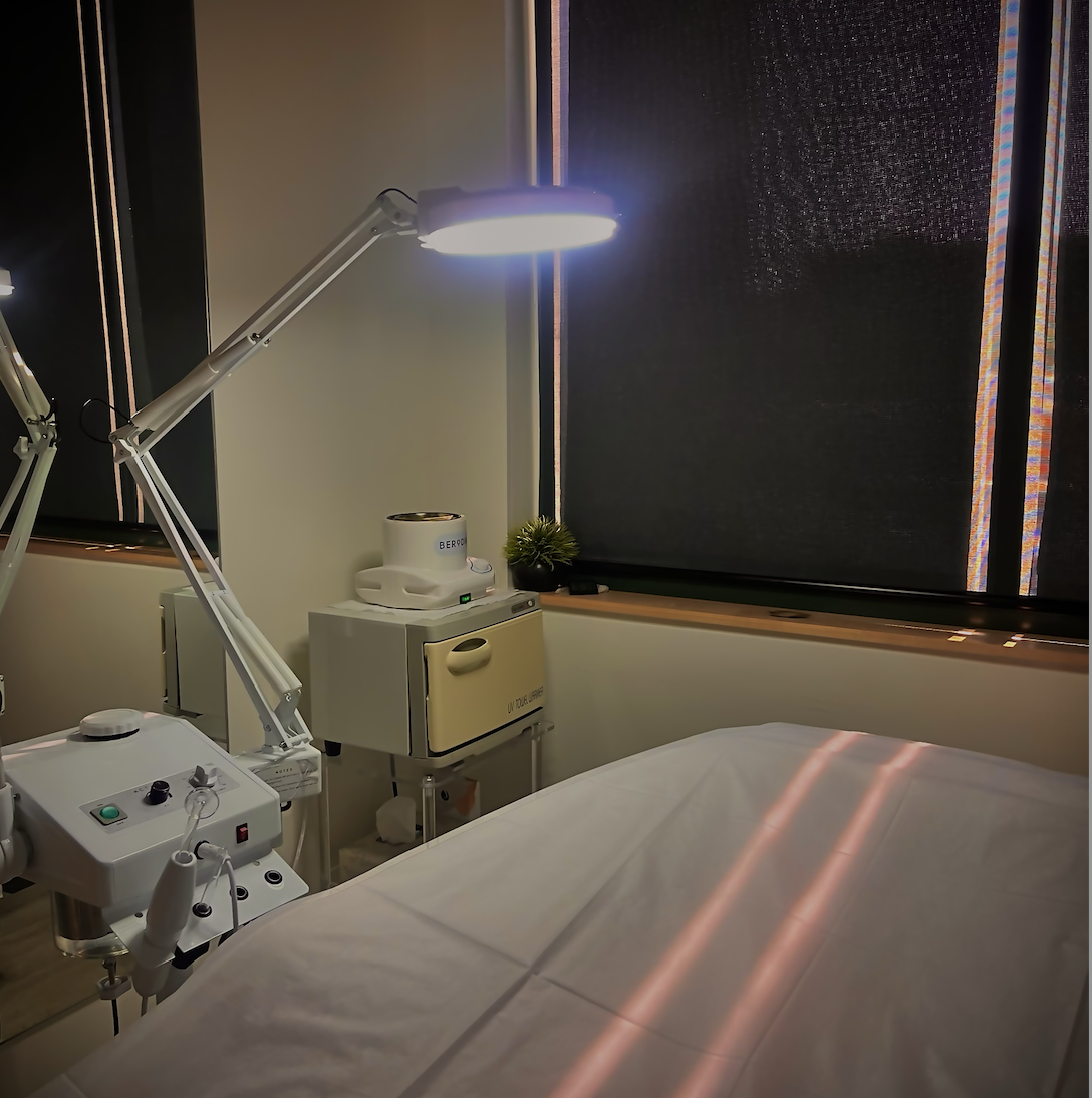 Medical examination room with a treatment bed, adjustable lamp, and medical equipment, with dark window blinds and a small potted plant.
