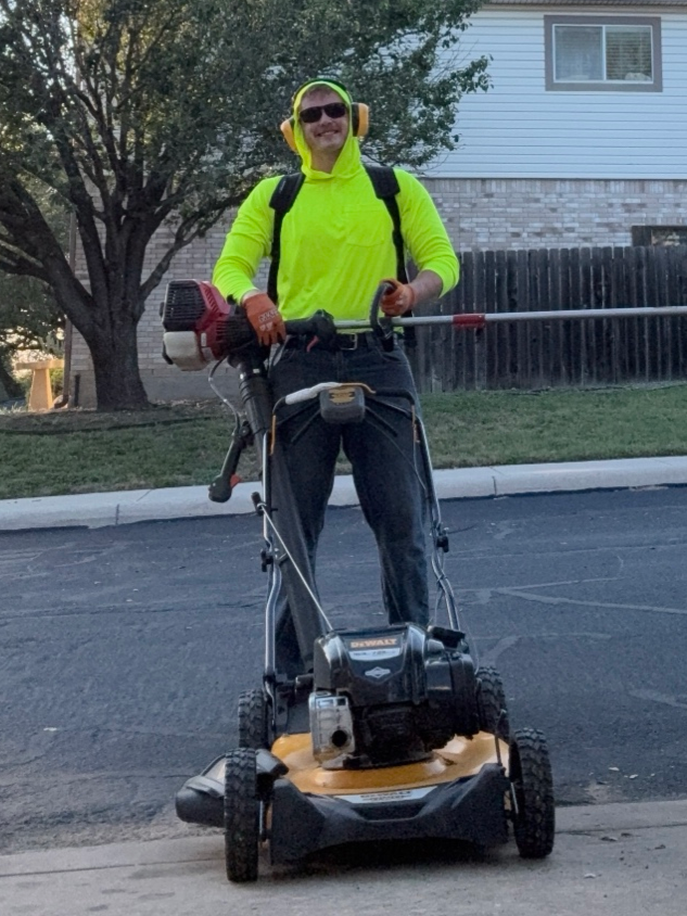 A man in a bright yellow shirt and sunglasses standing on a sidewalk with a string trimmer, and a mower.