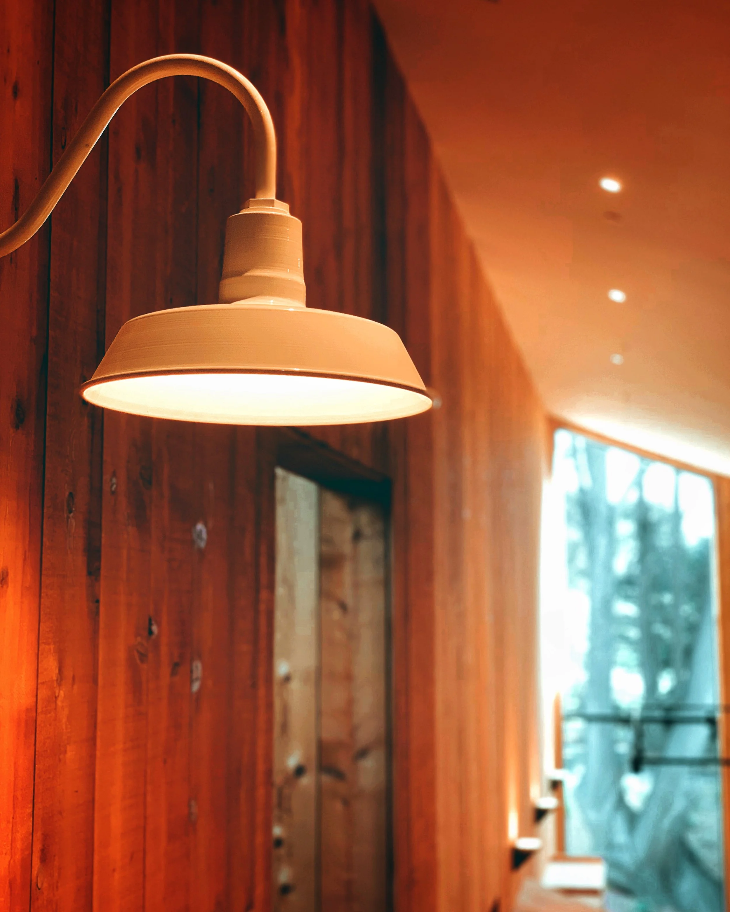 Close-up of a vintage white wall-mounted lamp with a rounded shade, attached to a wooden wall in a warm, cozy interior, with ceiling lights and window in the background.