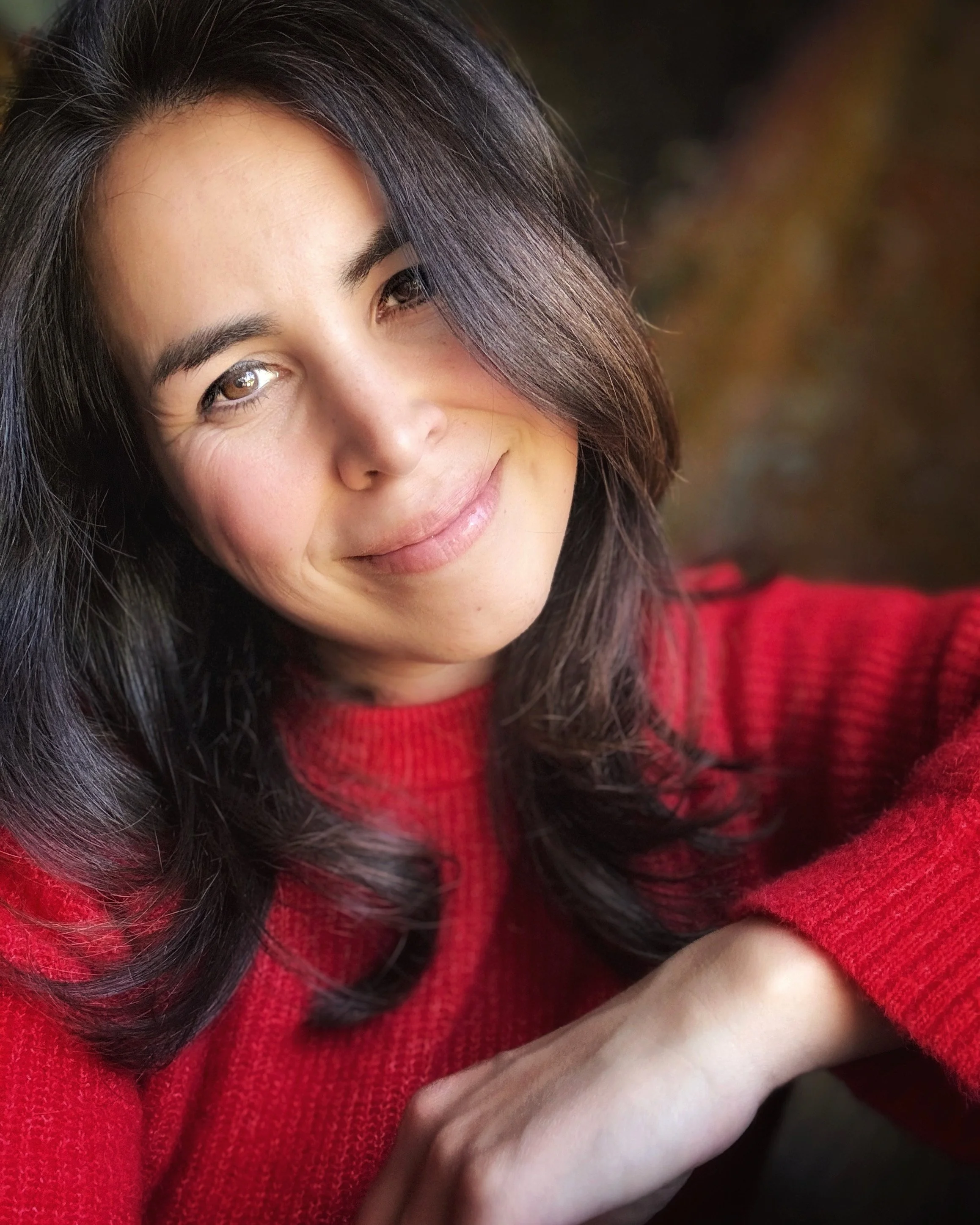 Close-up of a smiling woman with long dark hair wearing a red sweater, set against a blurred background.