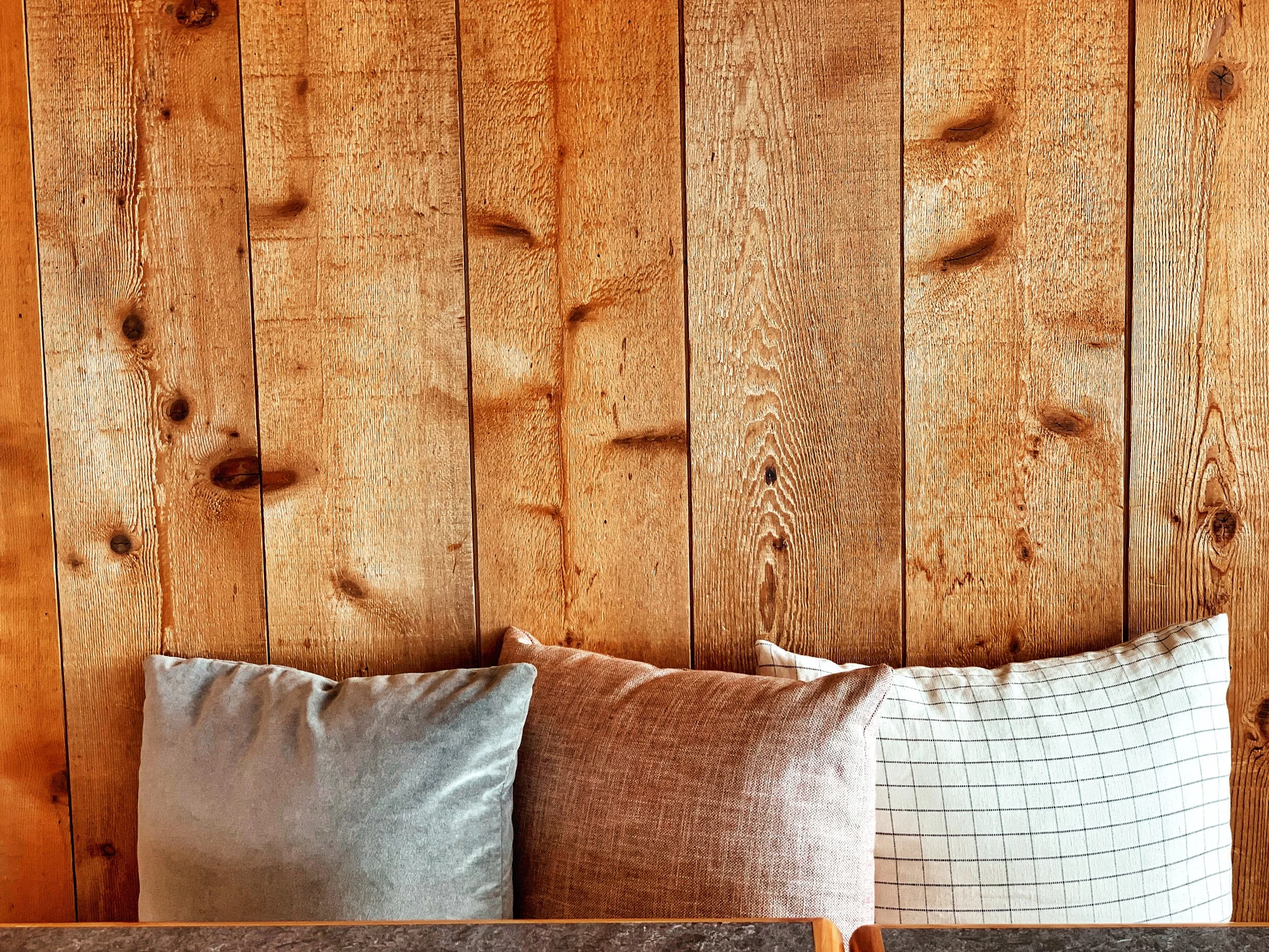 Three pillows on a bed against a wooden wall, with a gray pillow on the left, a reddish-brown pillow in the middle, and a checkered pillow on the right.