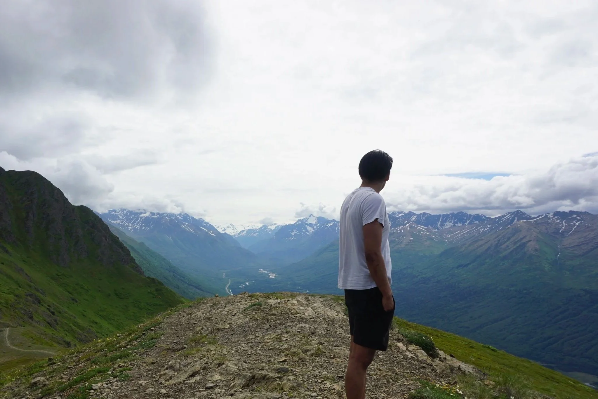 A man with dark hair and a white t-shirt standing on a mountain trail, looking at snow-capped mountains and green valleys under a cloudy sky.