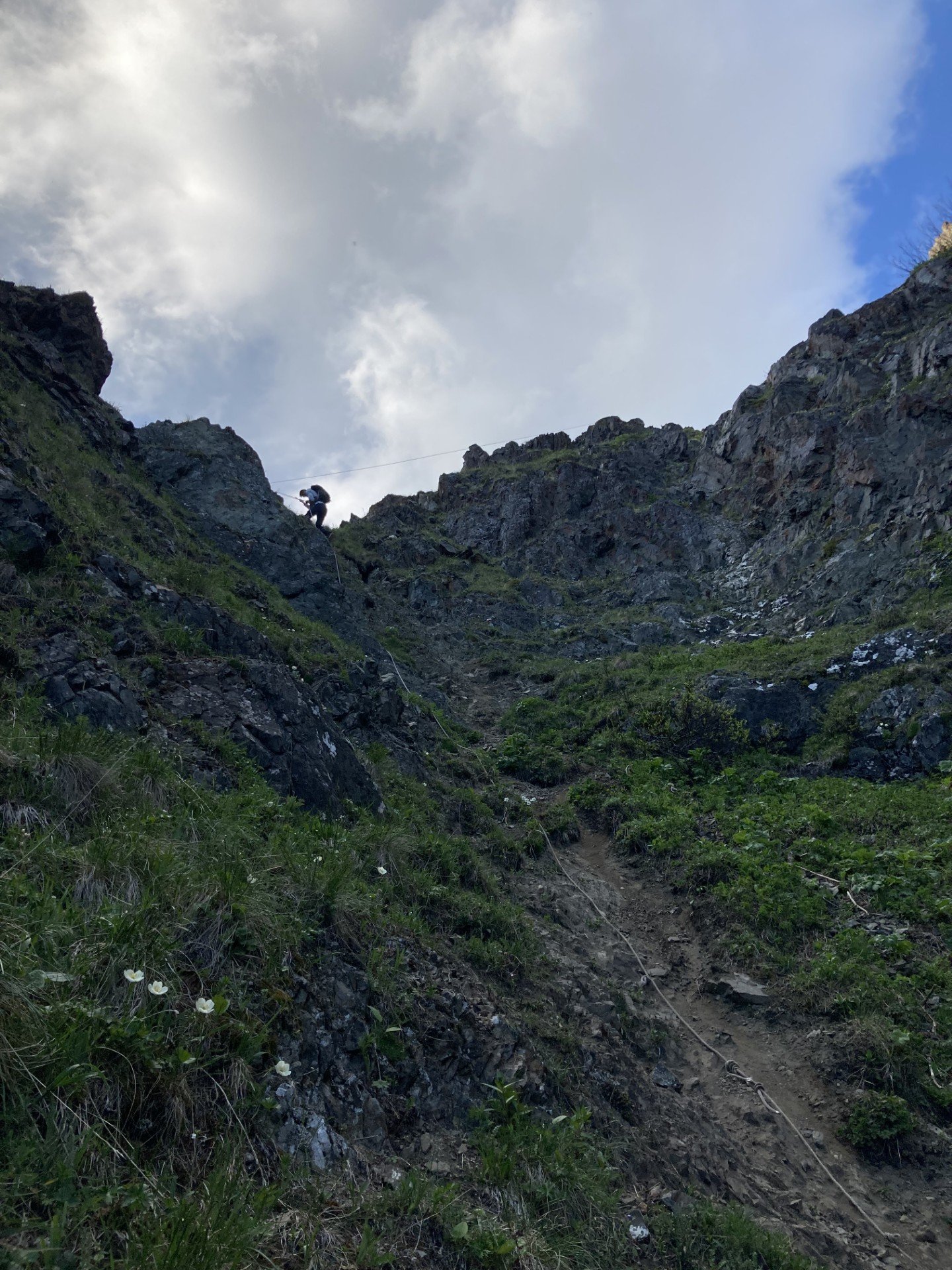 Person hiking on a steep rocky mountain trail with greenery, clouds, and blue sky in the background