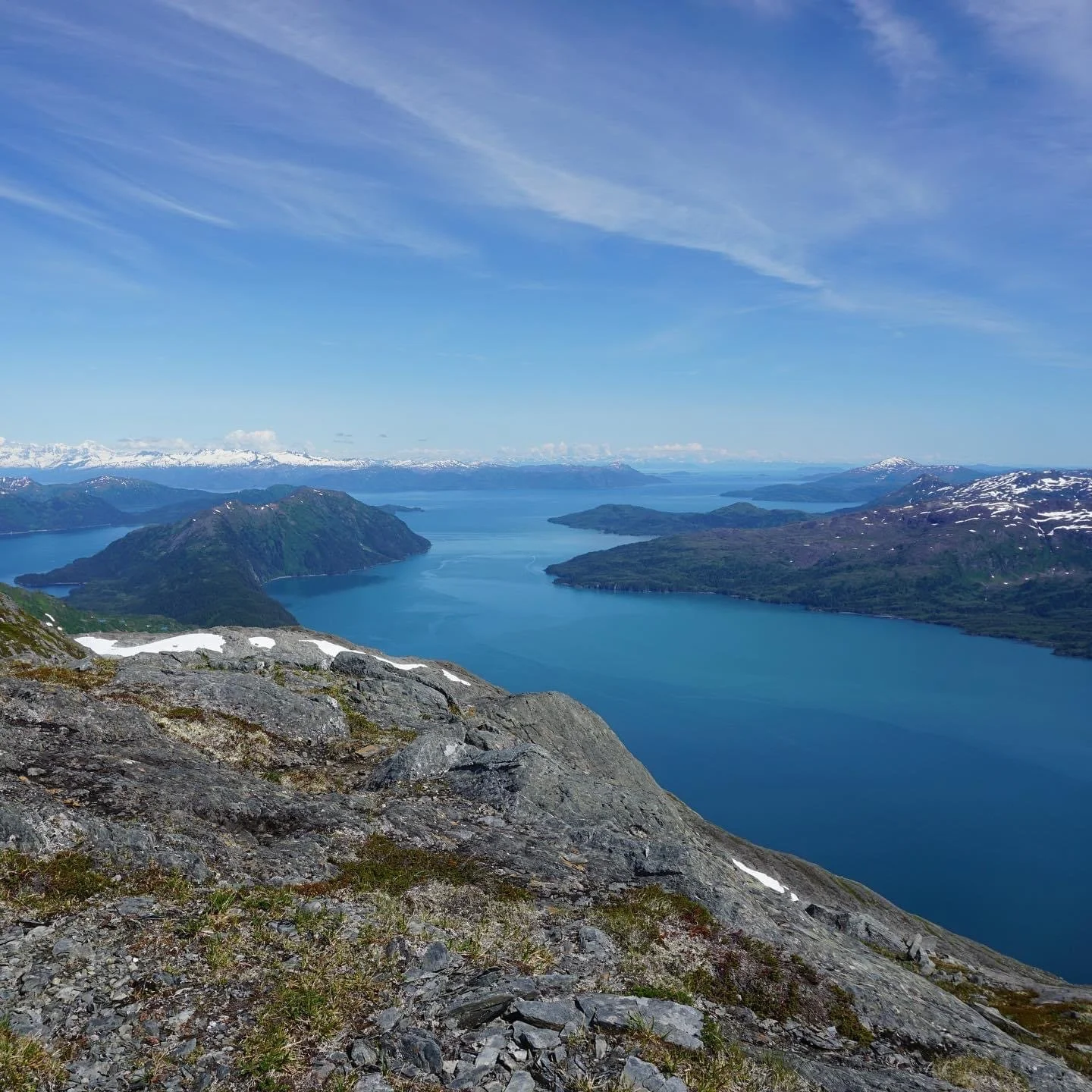 A panoramic view of a large, calm lake surrounded by mountains, some of which have patches of snow, with a partly cloudy sky above.