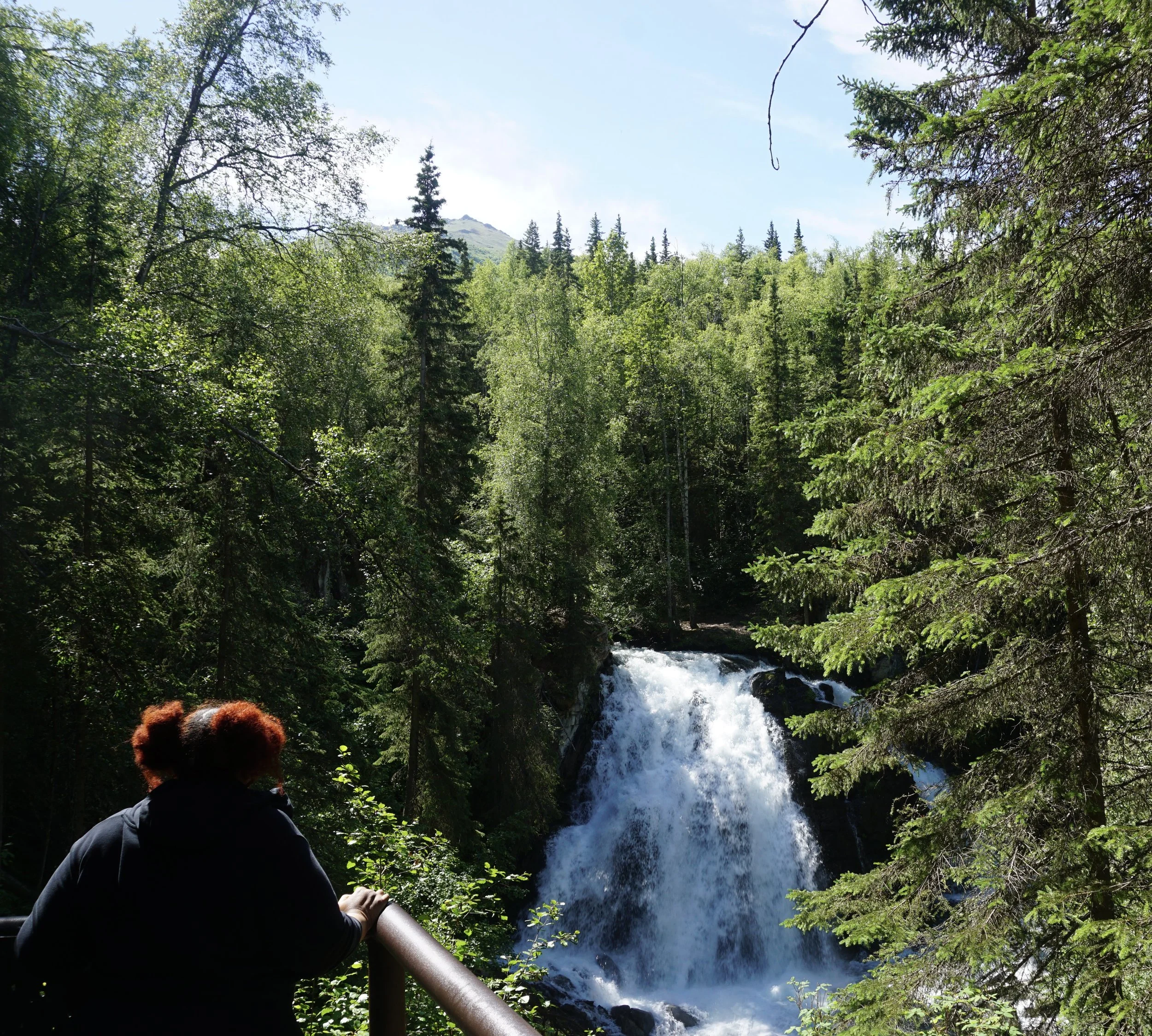 Person sitting by a waterfall in a lush green forest, with mountain and blue sky in the background.