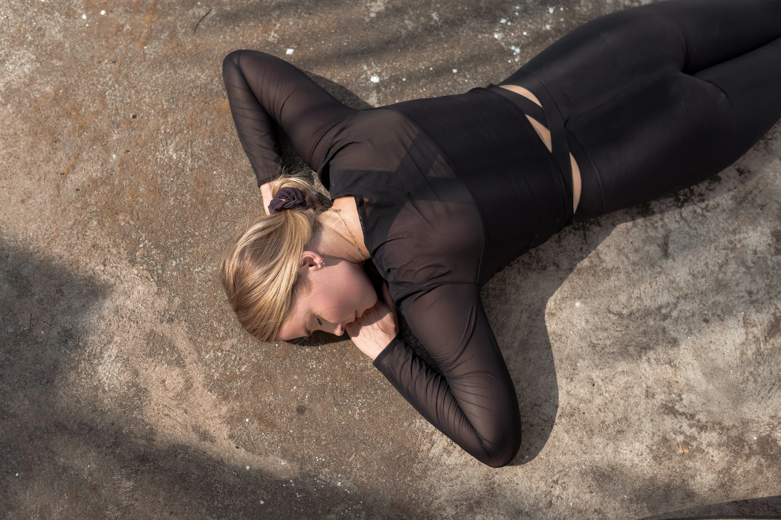 A woman with blonde hair, wearing black sheer long-sleeve top and black pants, lying on sandy ground with her arm behind her head, eyes closed.