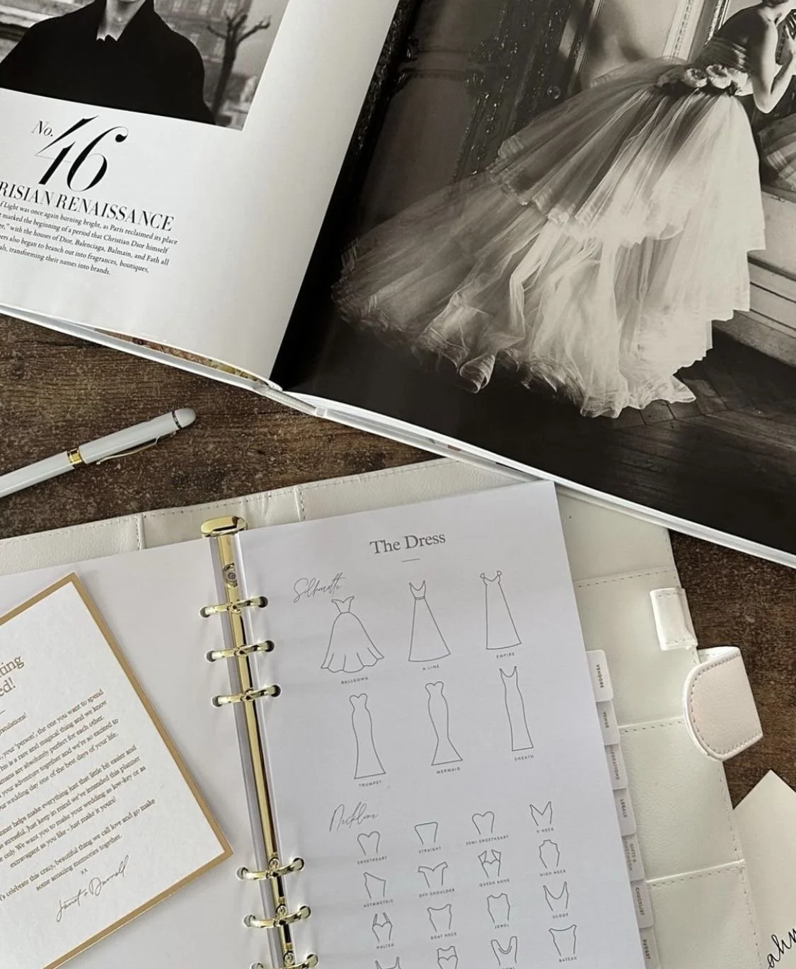 Open magazine showing black and white photos of a woman in a ball gown and a man in formal attire. A white pen, a ring-bound folder with fashion sketches of various dress styles, and a white planner are on a wooden table.