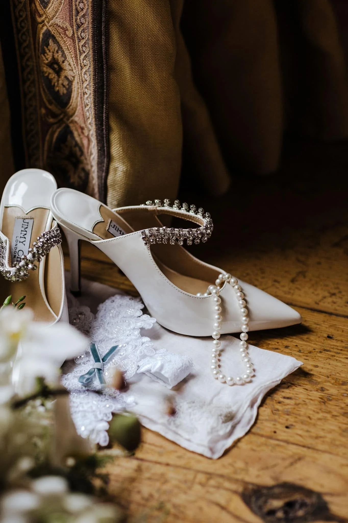 Close-up of a wedding shoe with pearl necklace and lace details placed on a white cloth, with a cushioned bench with dark scrollwork fabric in the background.