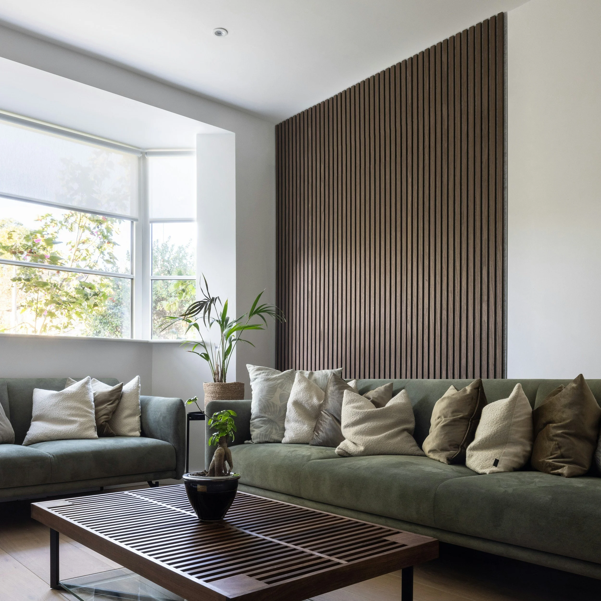 Modern living room with a green sofa, beige pillows, potted plant, wooden slat wall, large window with blinds, and a wooden slatted coffee table.