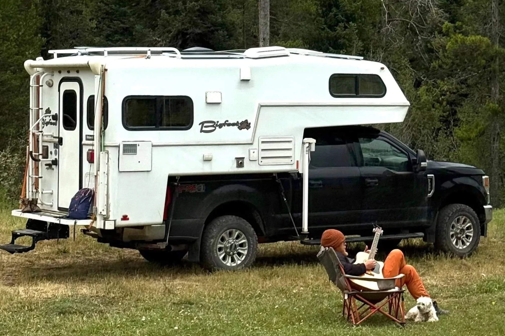 Kevin sits outside his home on wheels, playing guitar next to his dog, Jackson. A truck camper named Fred, sitting upon a grassy area surrounded by trees. Living life to the fullest, embodying his best self, and being present everyday.