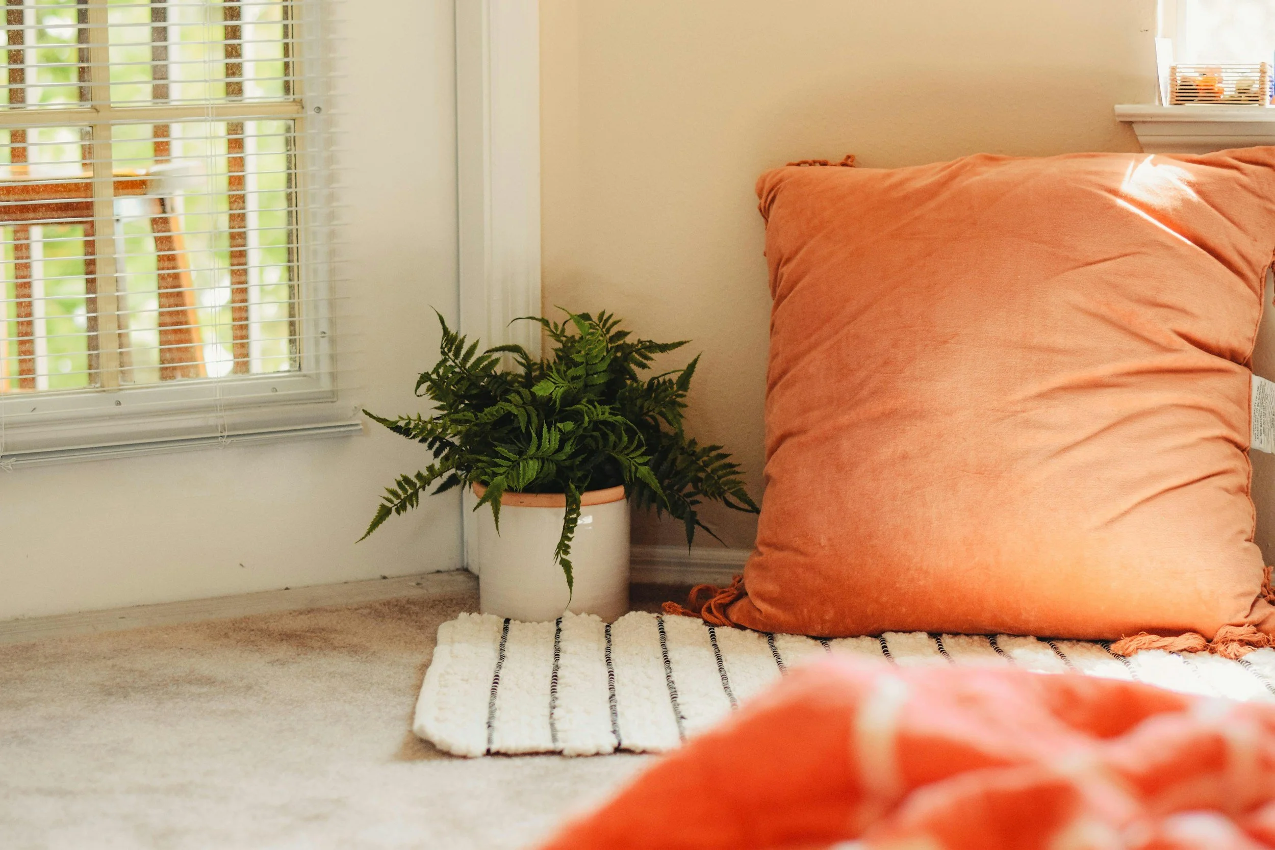 A corner of a room with a beige wall and a window with white blinds. There is a green potted fern on the floor, partially on a striped rug, next to an orange pillow on a woven mat.