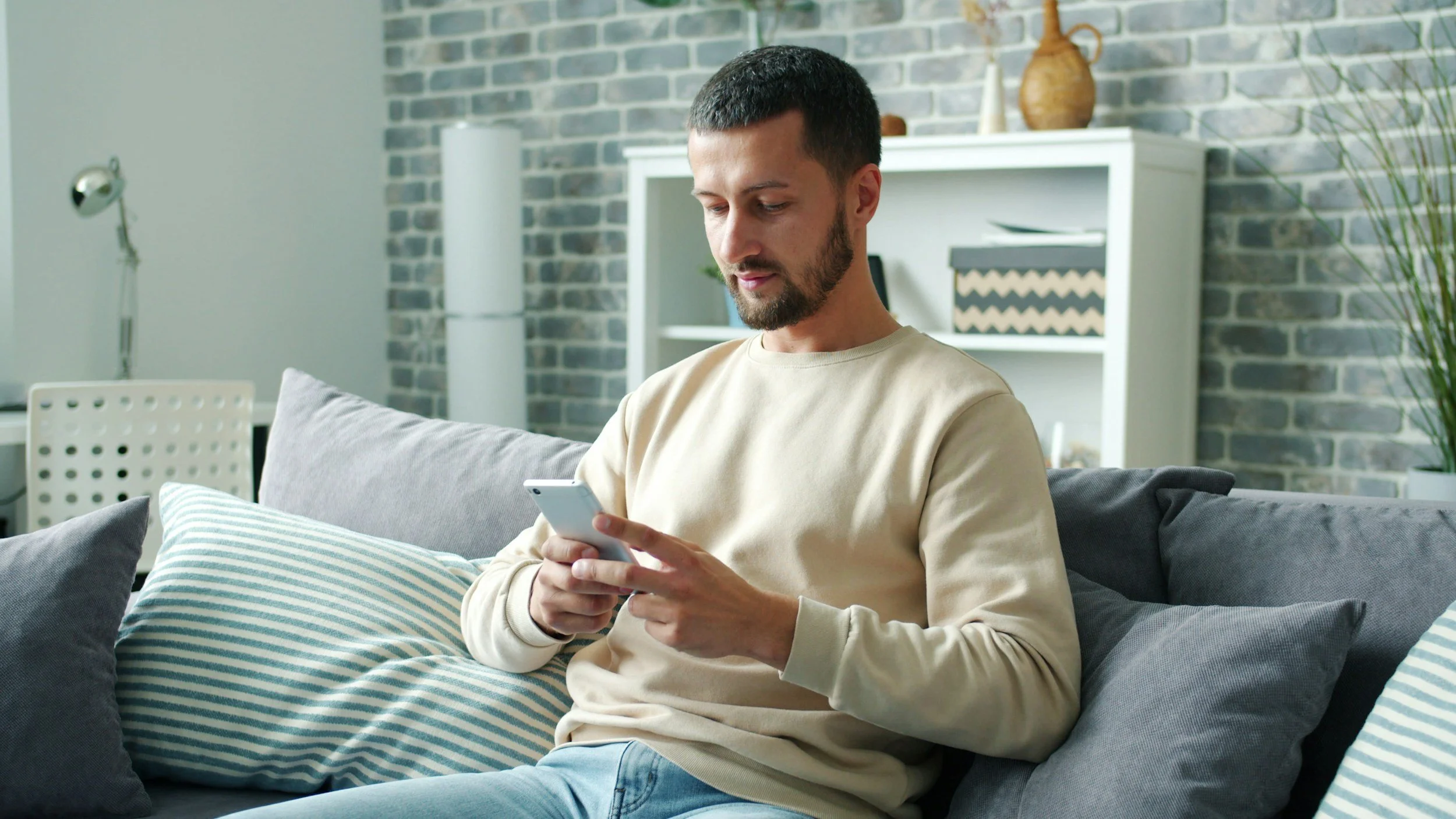 A man with short dark hair and beard sitting on a gray sofa, looking at his smartphone. The background features a brick wall, a white bookshelf, and decorative items.