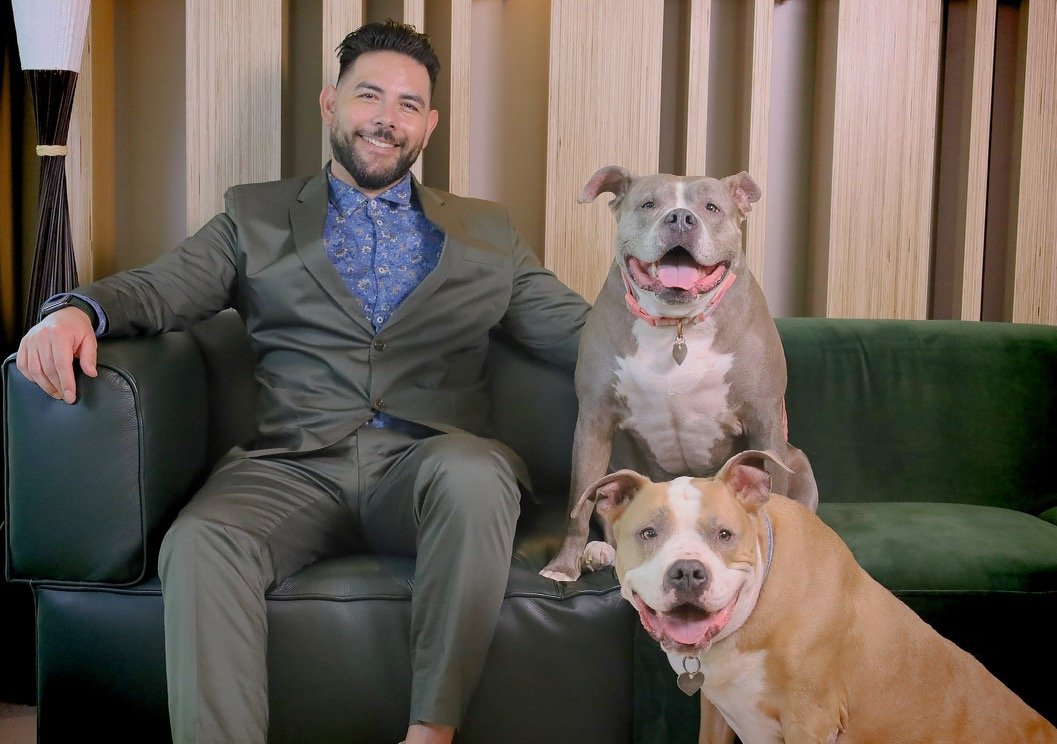 A man with a beard in a suit sitting on a green couch with two smiling dogs; one sitting on the couch and the other standing on the floor.