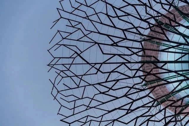 Close-up of a large, decorative outdoor art installation resembling a tree or branches, made of black metal rods against a partly cloudy sky.