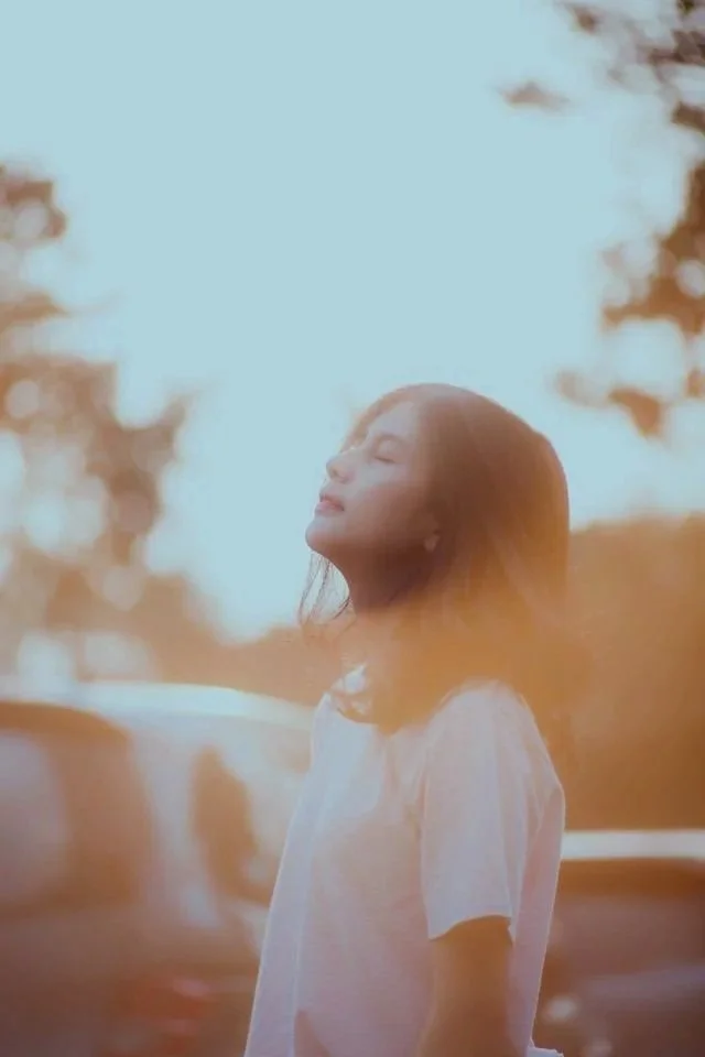 A young woman standing outdoors with her eyes closed and face turned upwards, bathed in warm sunlight with trees in the background.