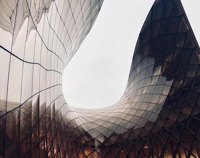 Modern architectural building with curved glass facade seen from below, reflecting the cloudy sky.