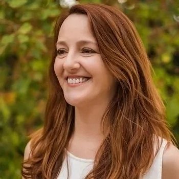 A smiling woman with long reddish-brown hair outdoors with green foliage in the background.