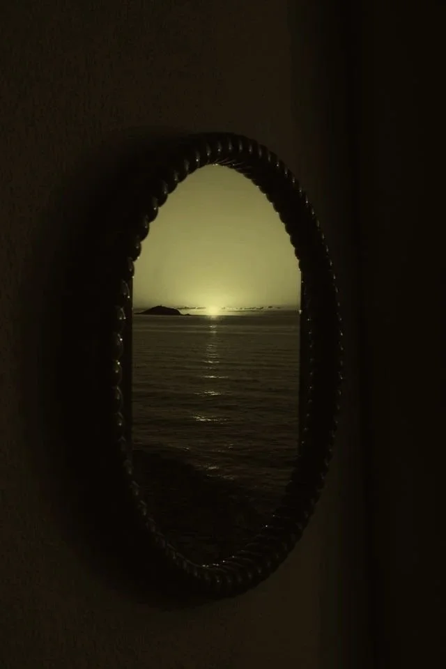 View of the ocean through an oval-shaped mirror illustrating a sunset with a small island in the distance.