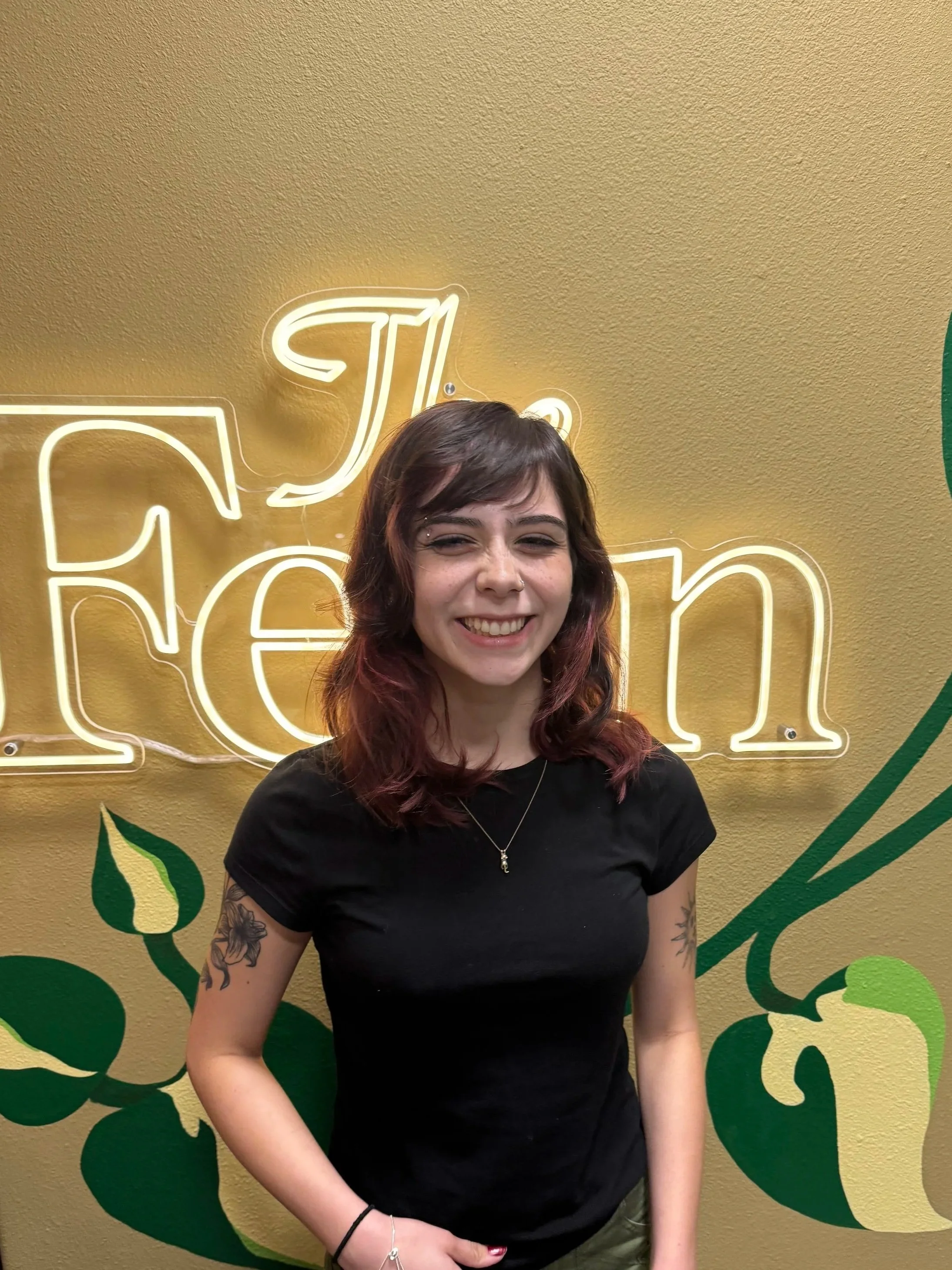 A woman with shoulder-length wavy hair, smiling, wearing a black T-shirt with tattoos on her left arm, standing in front of a yellow wall with neon signage that reads 'The Fern', with green decorative leaves.