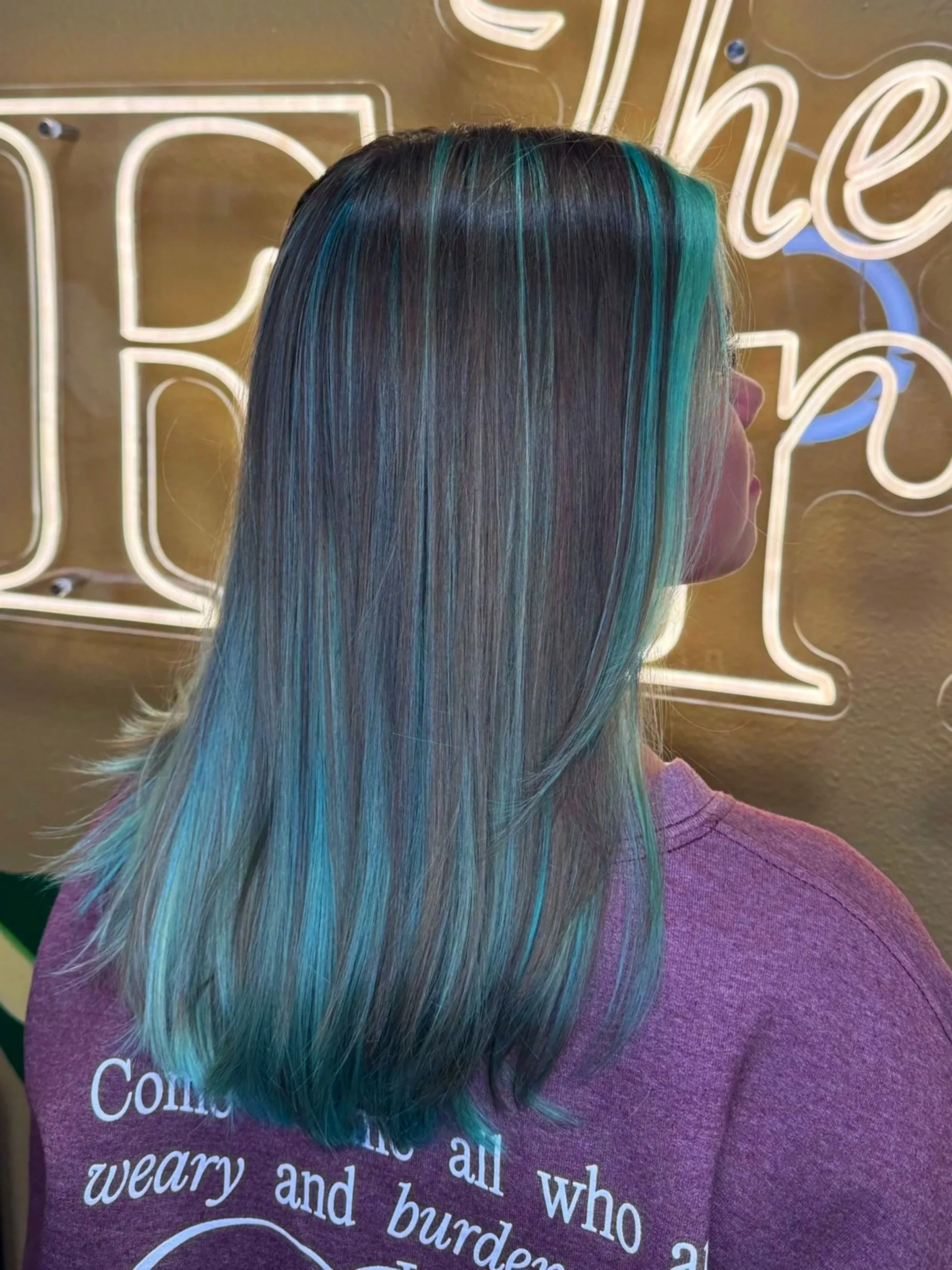A woman with straight, shiny hair dyed in shades of black, teal, and blue, standing in front of a neon sign with cursive writing. She is wearing a purple top with white text.