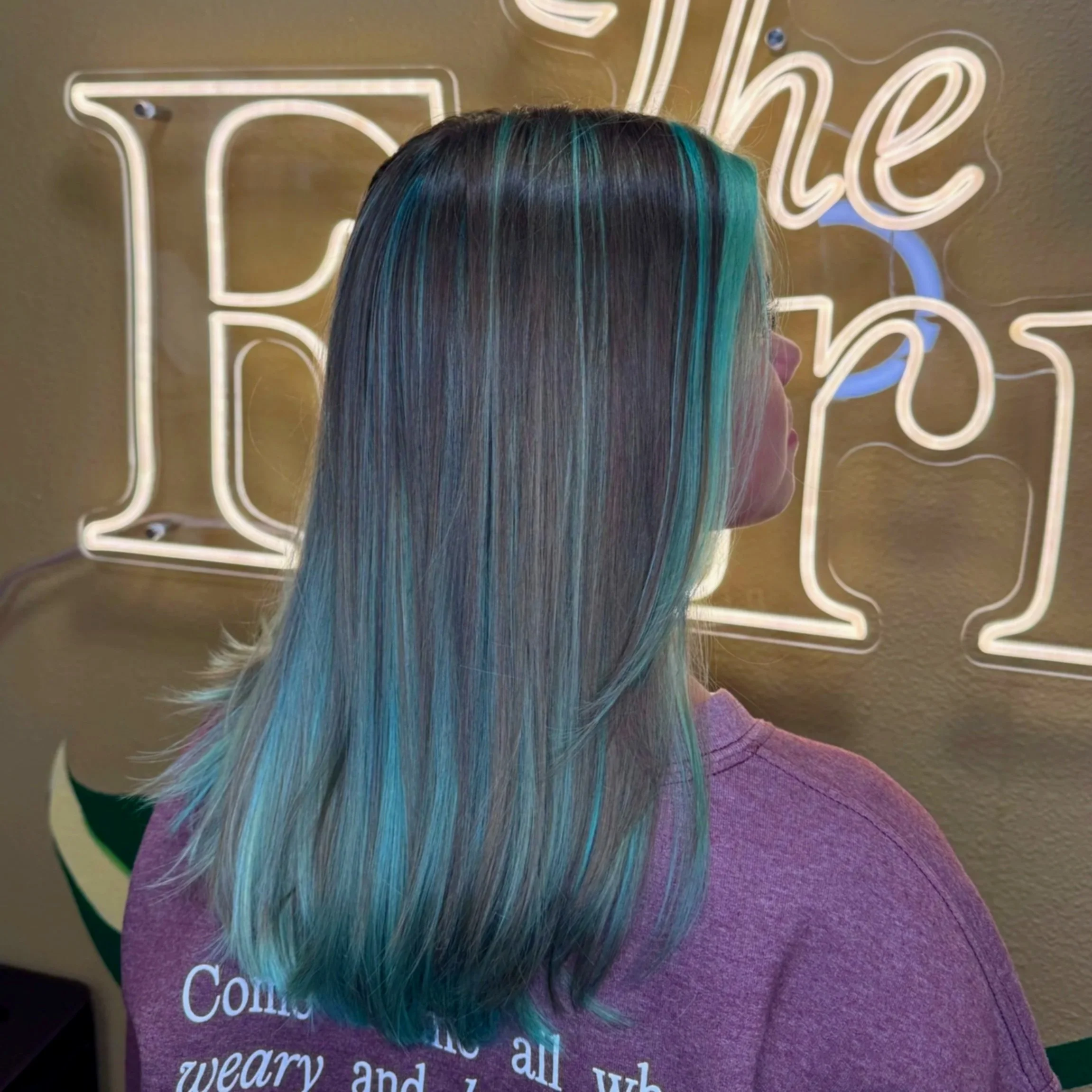 Back of a person with long hair dyed with shades of blue, green, and purple, standing in front of a neon sign with the word 'The Fern' partially visible.