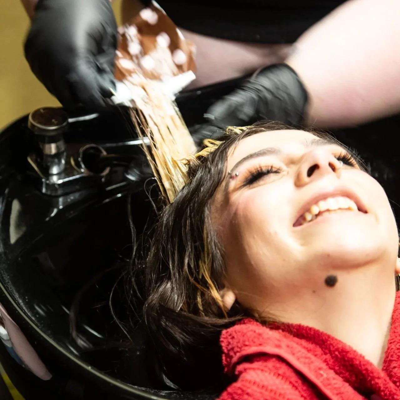 Woman with dark hair receiving a hair wash at a salon, lying back with her eyes closed and smiling.