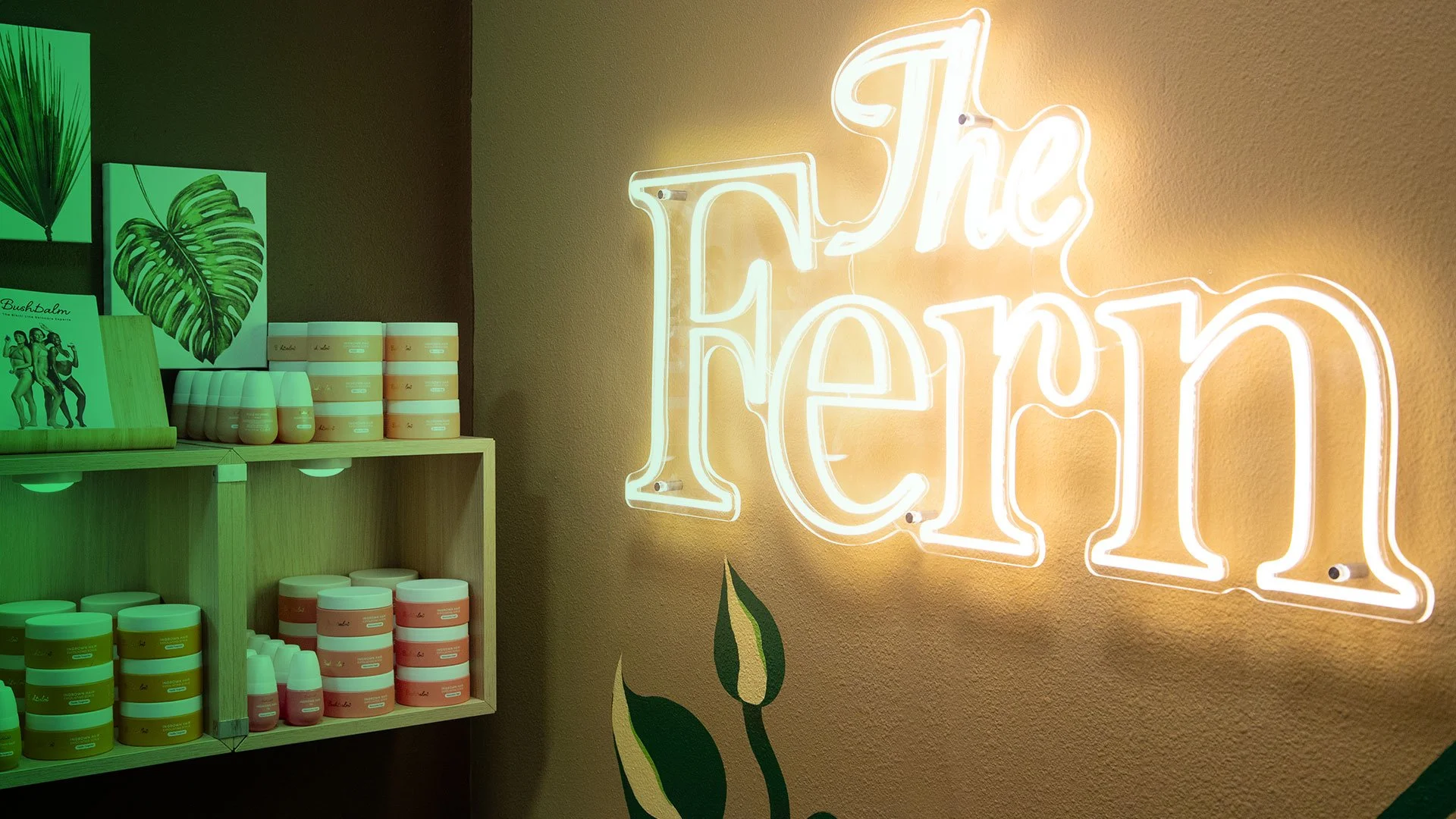 Neon sign that says 'The Fern' on a beige wall with part of a green plant illustration and shelves with jars of creams or lotions.