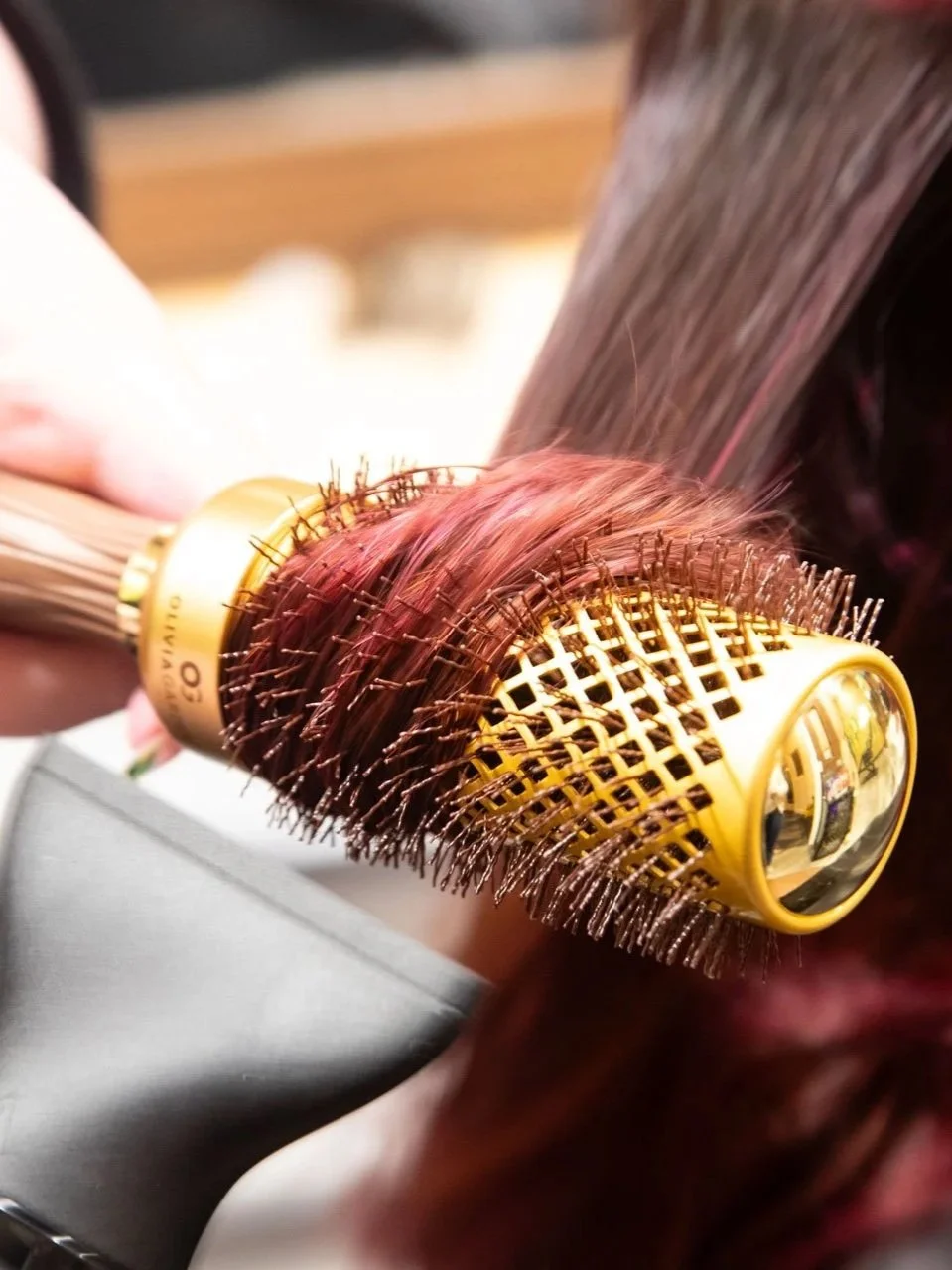 A person using a yellow round hairbrush with airflow vents to style or dry hair, with pink-colored hair strands visible.