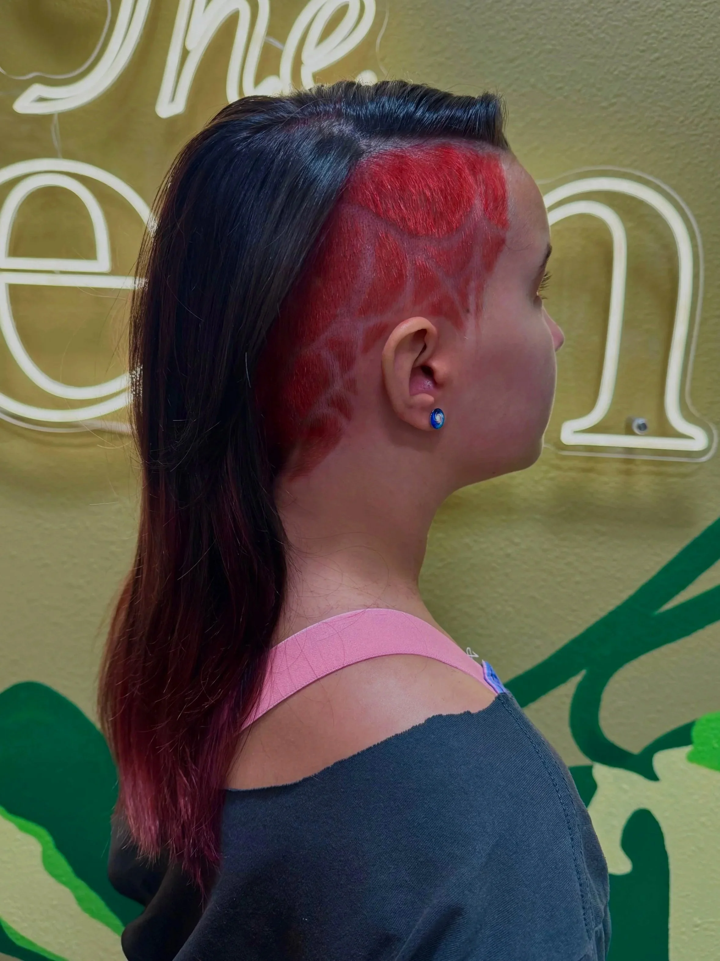 Side profile of a woman with long dark hair that transitions to red towards the ends, standing against a yellow-green background with decorative white and green design elements, wearing a pink top and a blue earring.