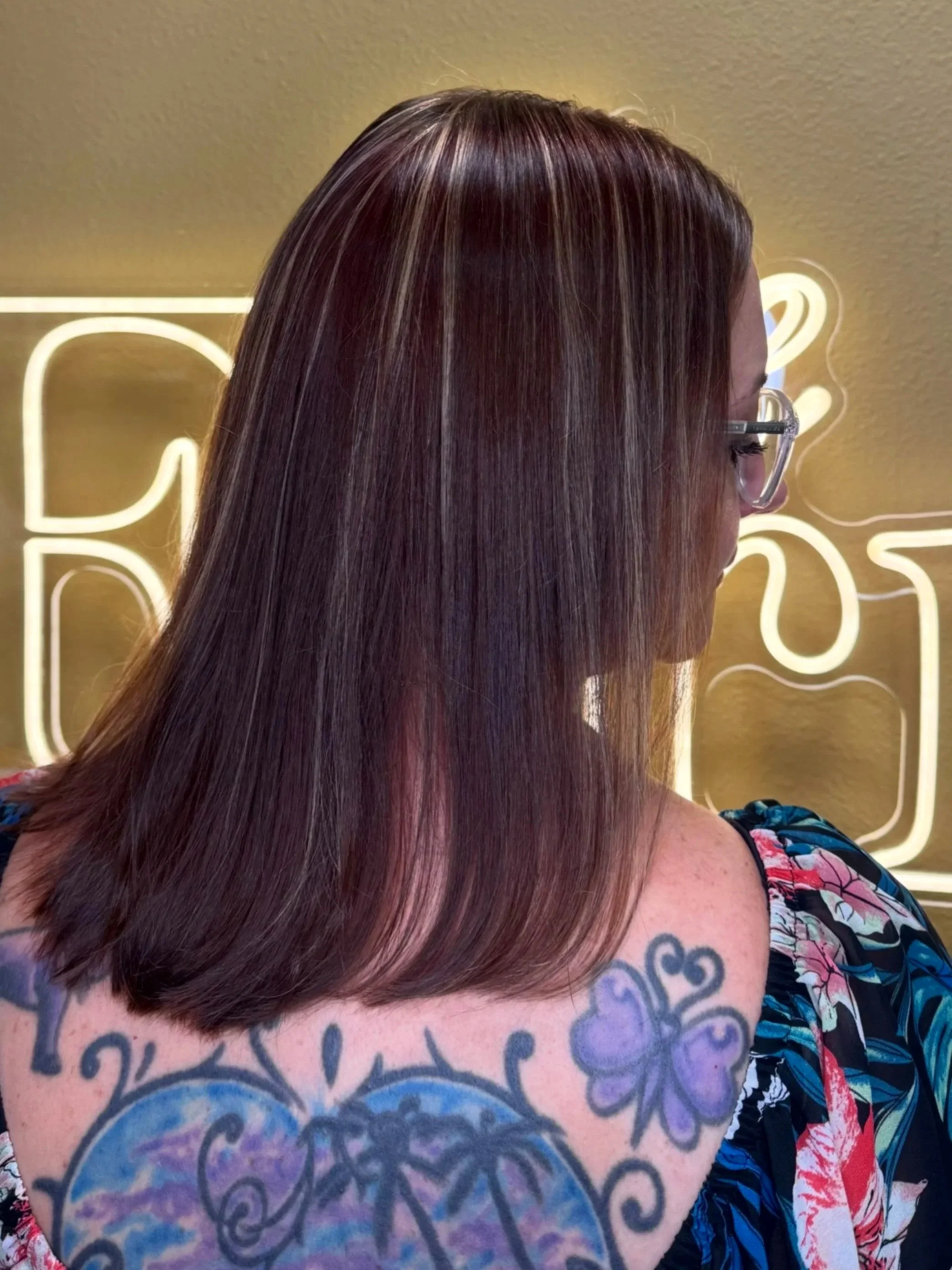 Side portrait of a woman with straight, layered, brown hair with blonde highlights, wearing glasses, and showing a colorful tattoo on her shoulder with floral and swirling patterns, wearing a floral dress, standing in front of a neon sign.
