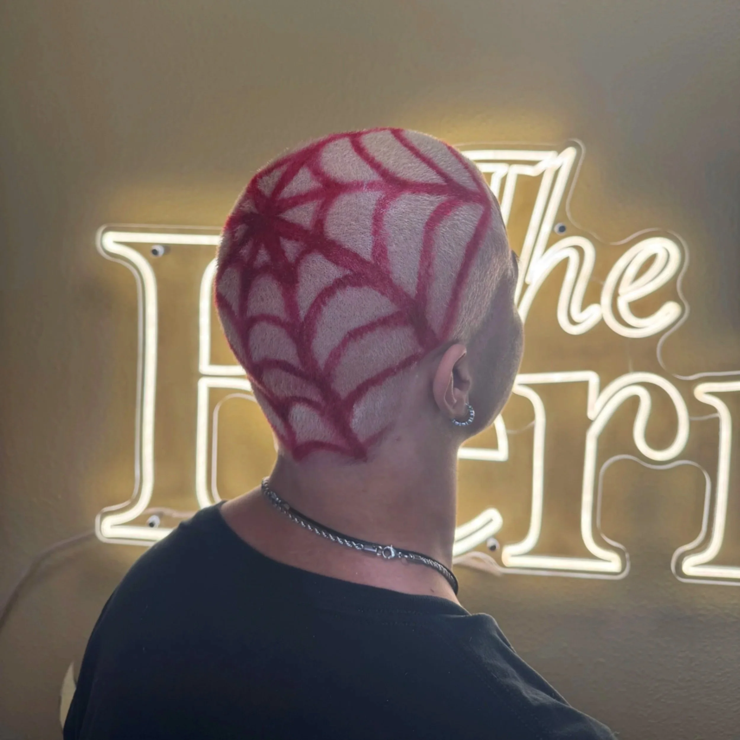 Close-up of a person with a red spider web pattern design on short hair, black shirt, chain necklace, and a bracelet, with a neon sign in the background that reads 'The Fern'.